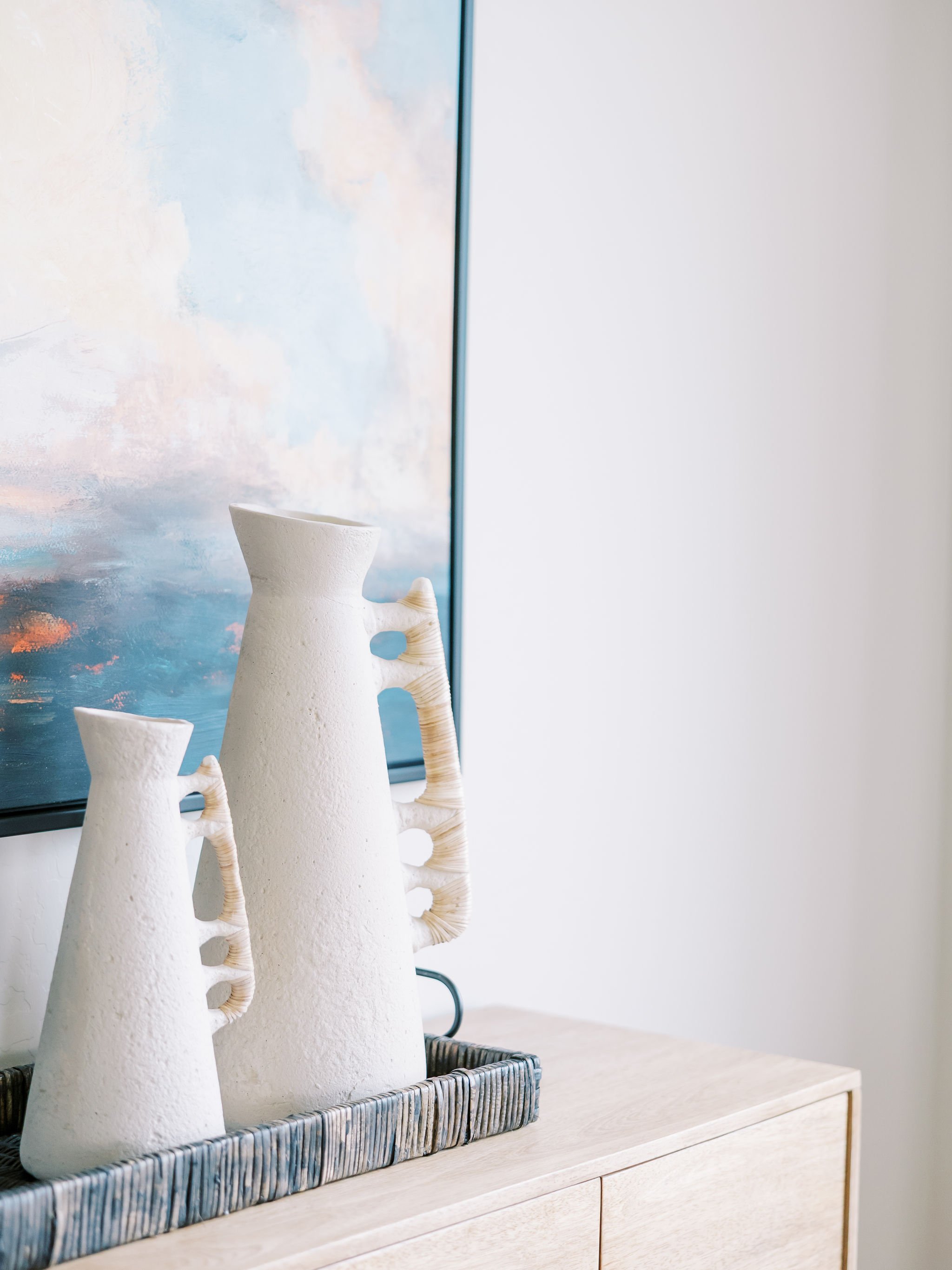 Two white textured ceramic vases with curved handles sit in a black wire tray on a light wood surface, with a colorful abstract painting in the background.