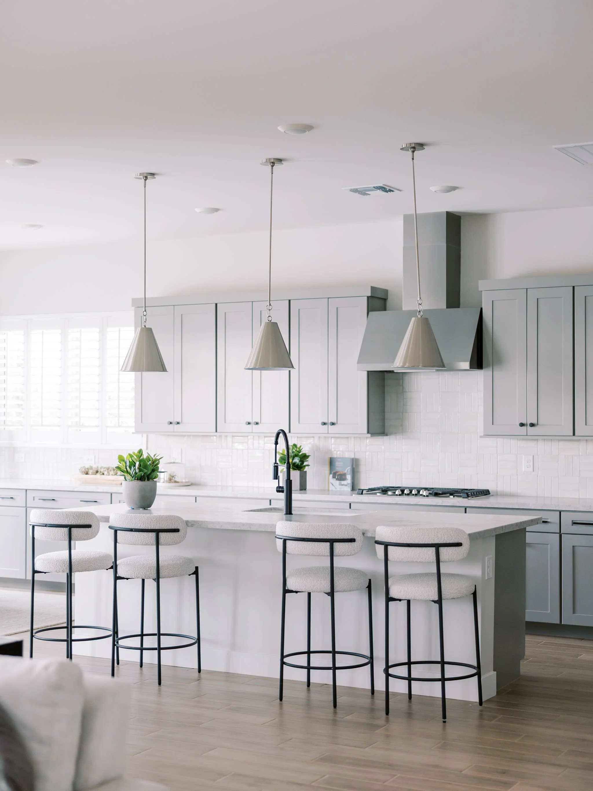 Modern kitchen with gray cabinets, white countertops, a white backsplash, three hanging pendant lights, a black faucet, bar stools with white cushions and black legs, potted plants, and a window with white shutters.