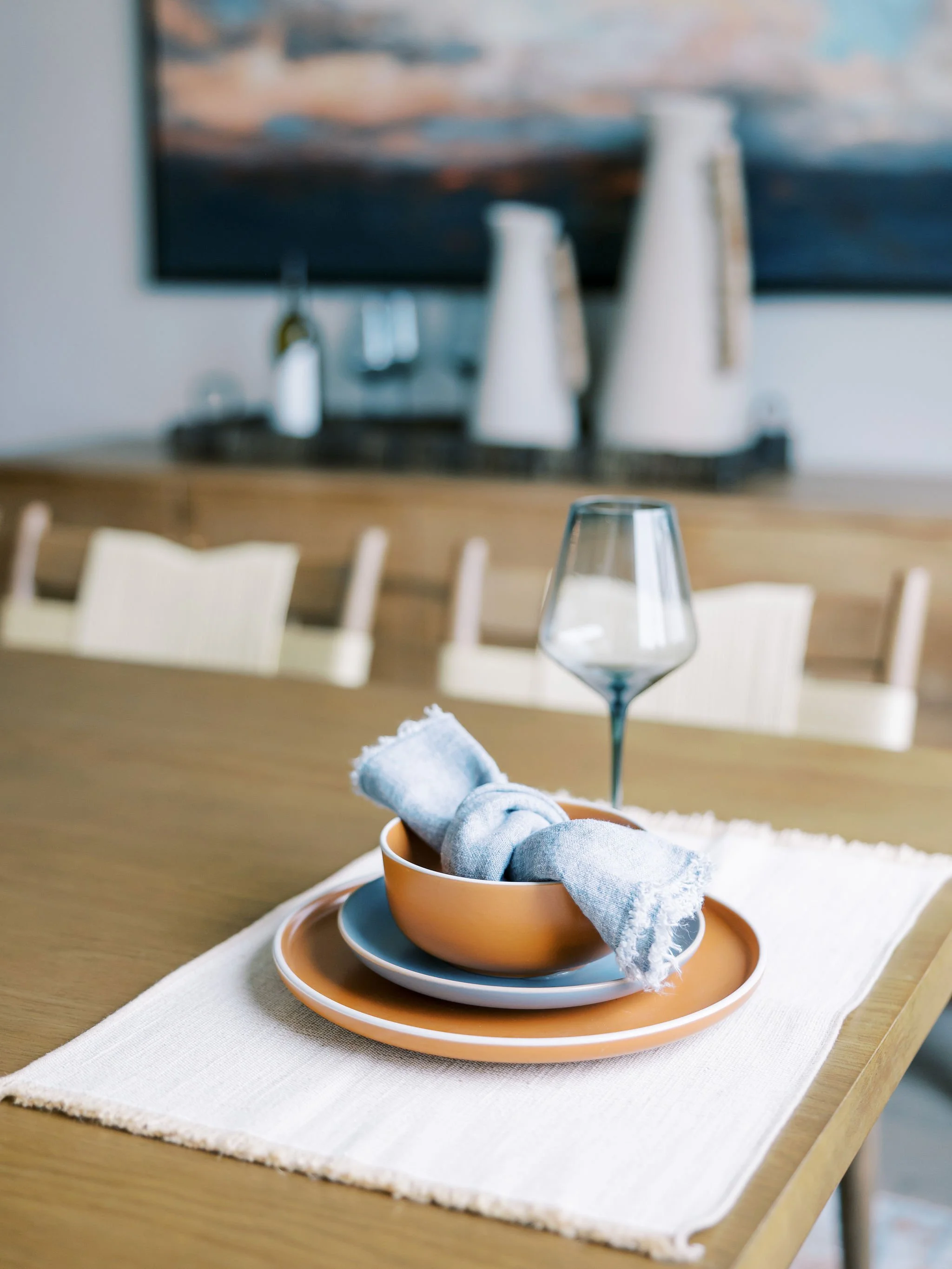 A dining table with a white cloth, set with a white wine glass and a stacked set of beige and white plates with a neatly folded light gray napkin inside a beige bowl, in a well-lit room decorated with modern art and neutral-colored furniture.