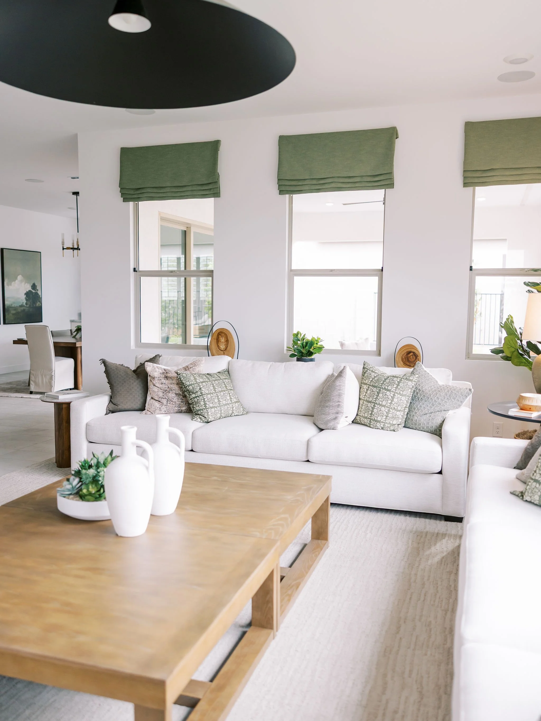 Bright living room with a white sofa, patterned throw pillows, a wooden coffee table with decorative vases and plants, and three windows with green Roman shades.