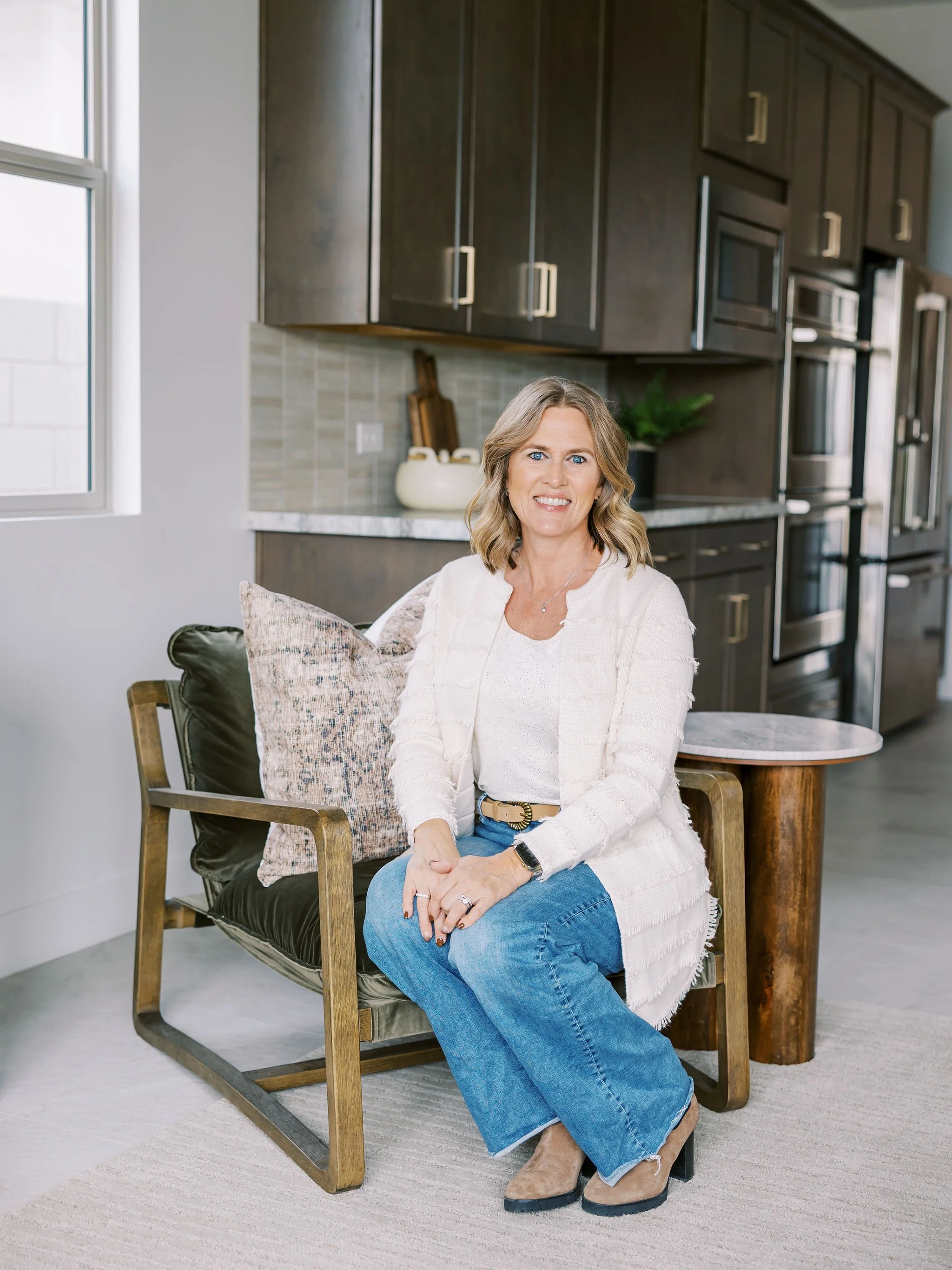 Jessica Curtis sitting on a wooden chair in a modern kitchen, smiling at the camera, with dark cabinets, a window, and a countertop with decorative items in the background.
