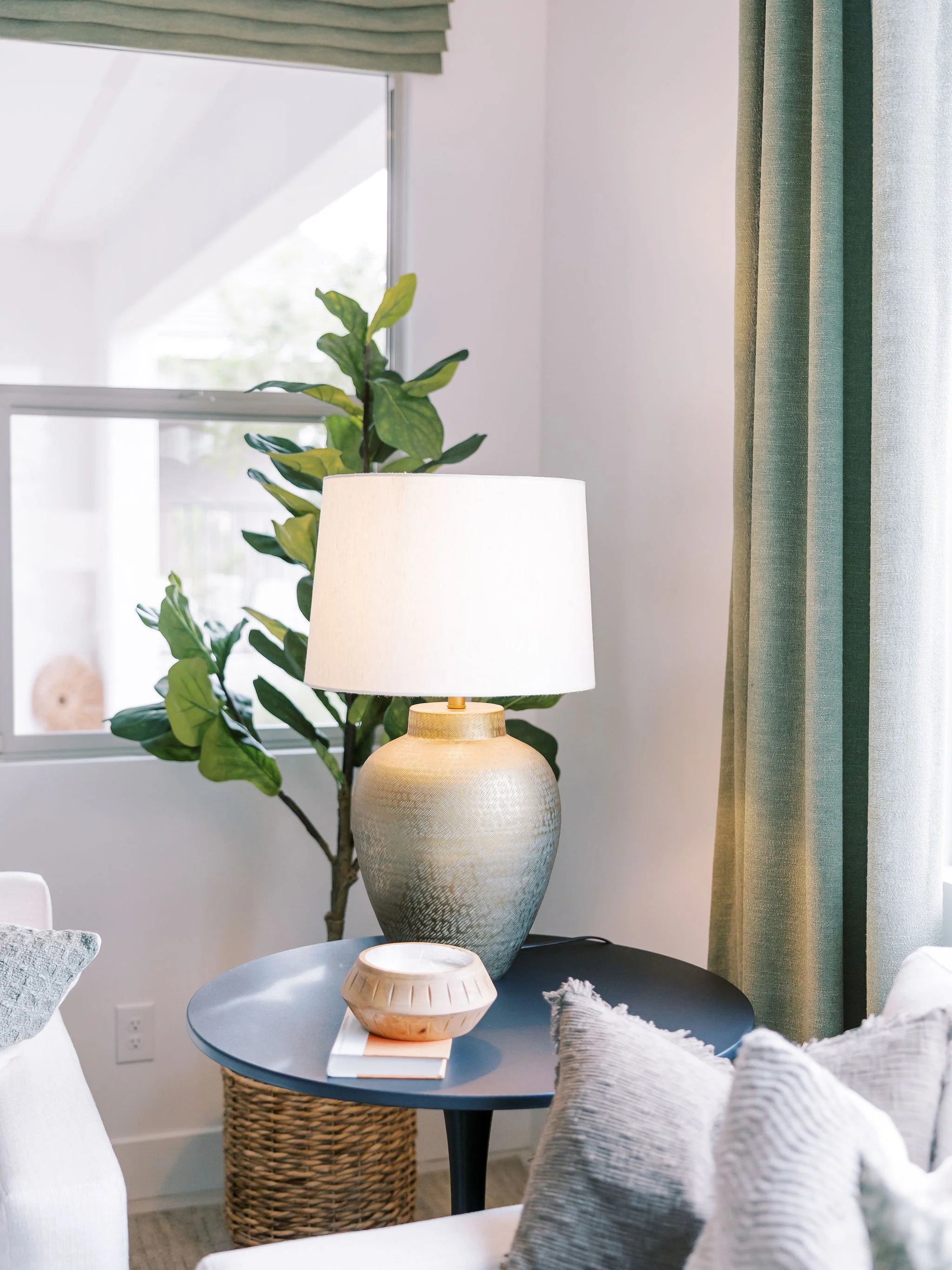 A living room side table with a large ceramic lamp, a decorative bowl, and a closed book, next to a sofa with textured pillows, a window with green curtains, and a tall leafy plant in the background.