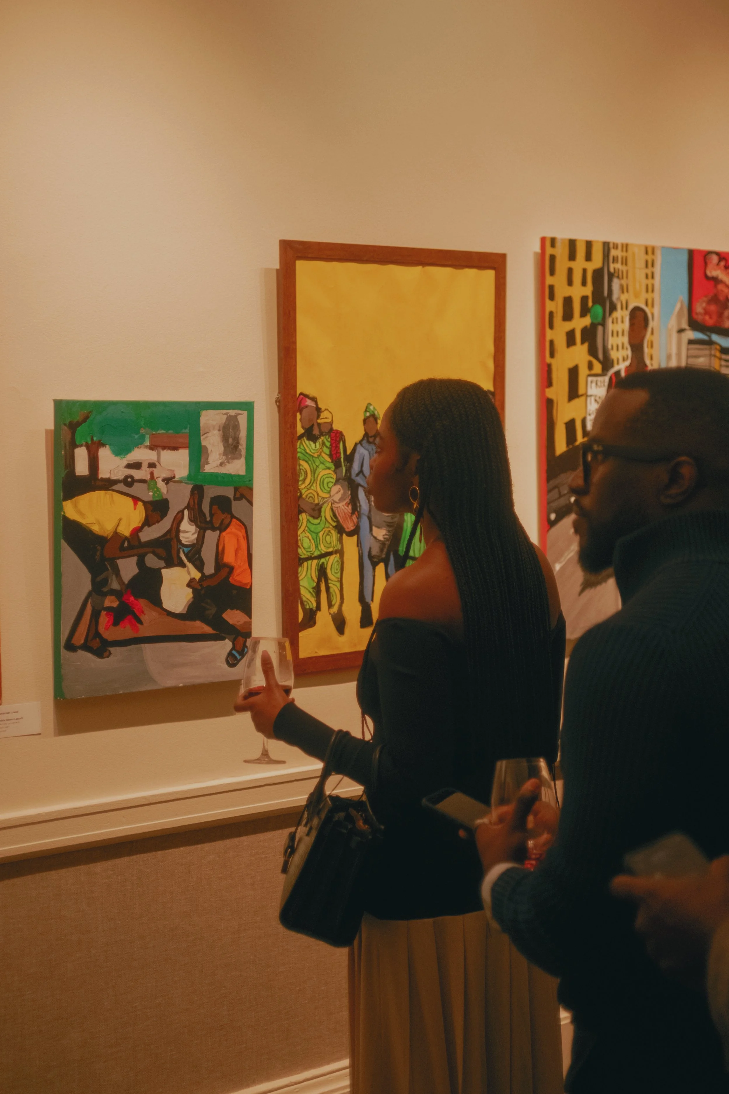 A woman with long braided hair and a man with glasses are observing colorful paintings in an art gallery while holding glasses of wine.
