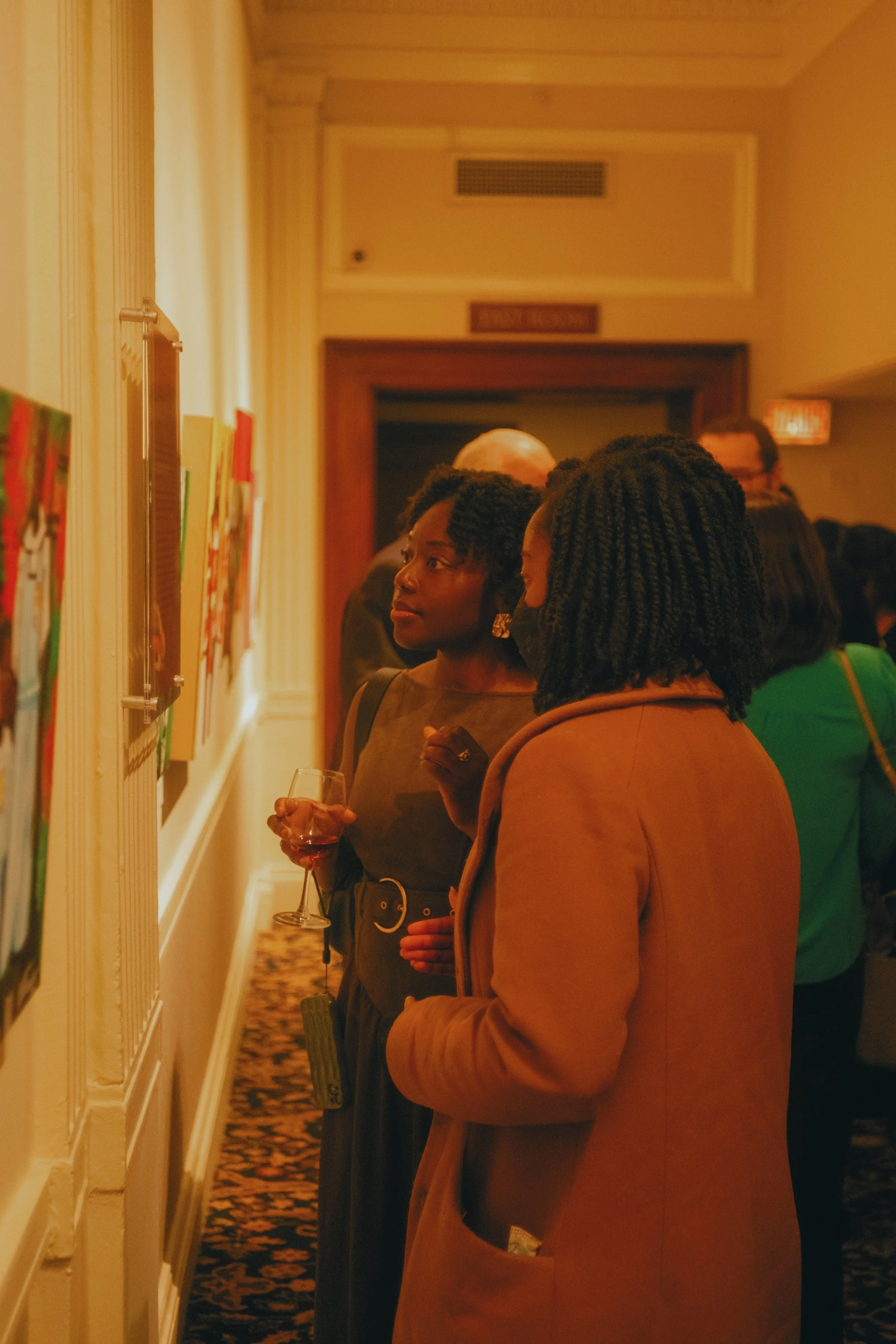 Two women looking at art displayed on a wall at an art gallery or exhibit, with one holding a glass of wine.
