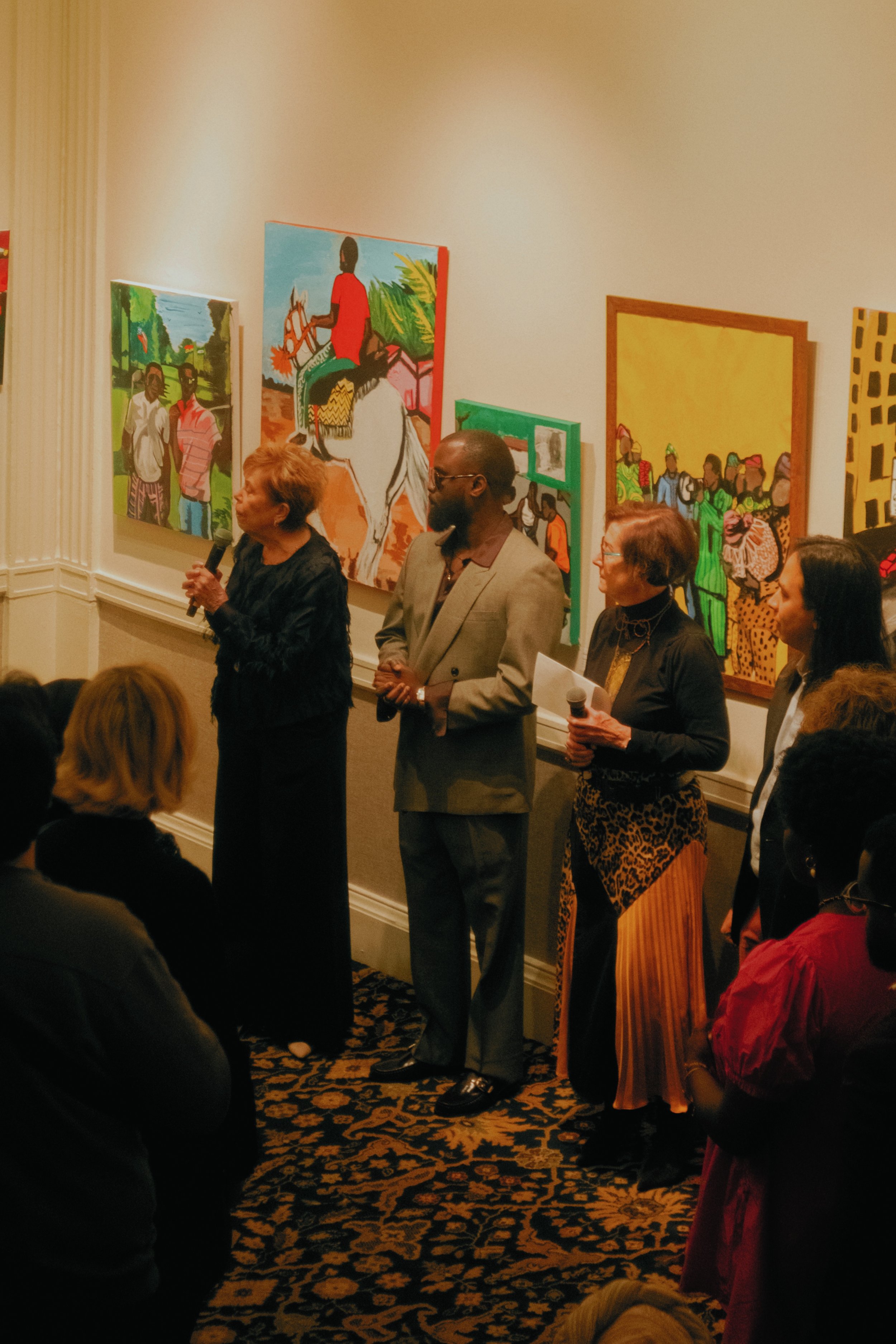 People standing in front of colorful paintings during an art gallery event, with some holding microphones and notes.
