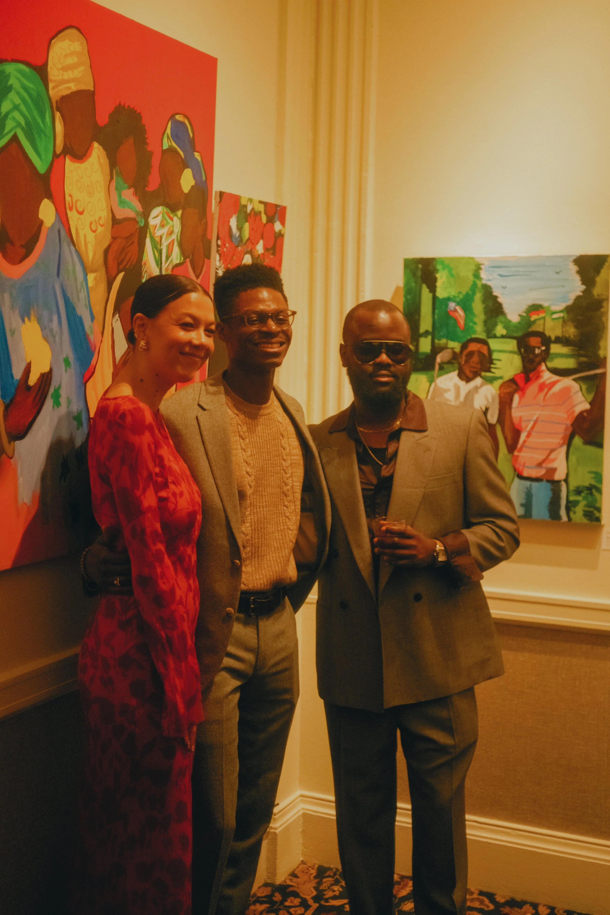 Three people standing in an art gallery in front of colorful paintings, dressed in formal and semi-formal attire, with two men wearing sunglasses.