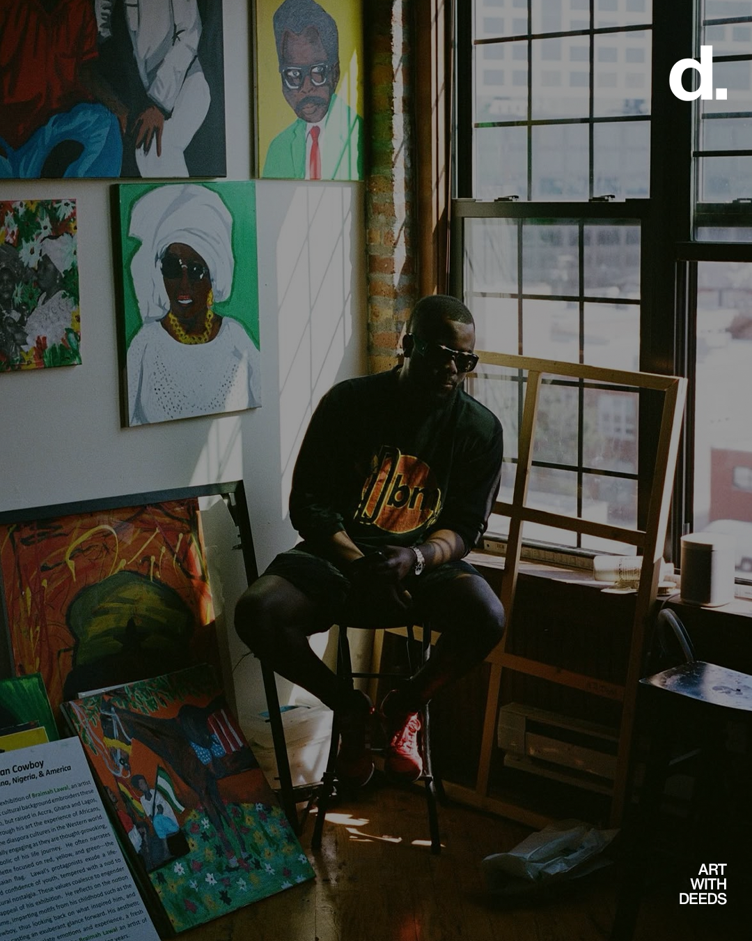 A man wearing sunglasses, a black jacket with a colorful logo, and red shoes, sitting on a stool in an art gallery with paintings on the wall and an open window letting in natural light.