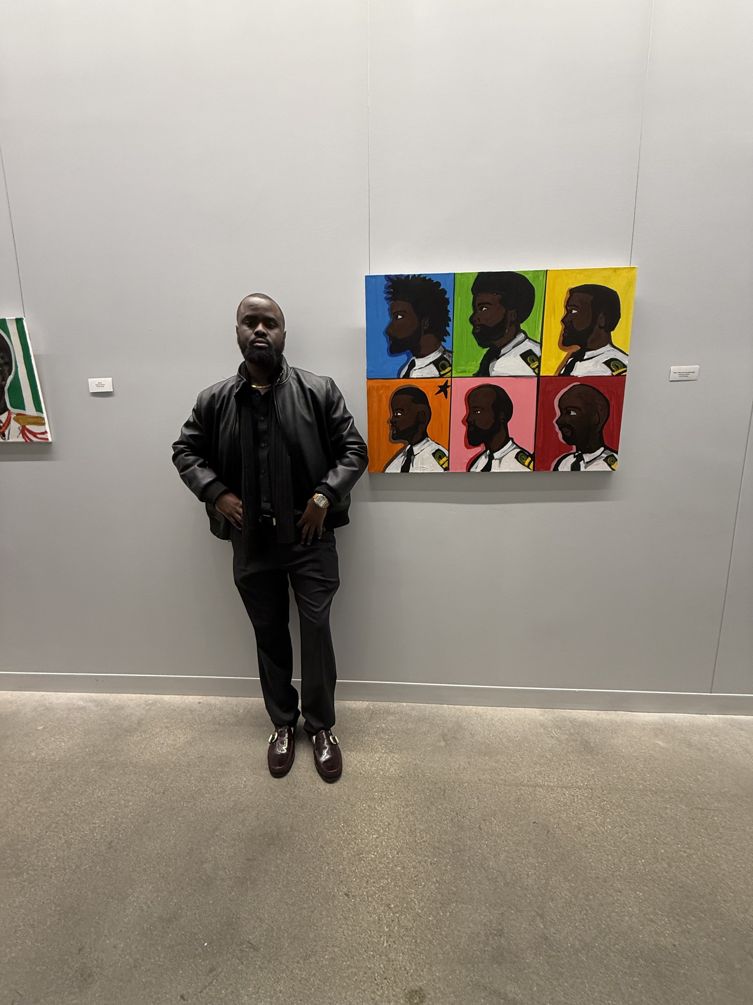 Man in black leather jacket standing in an art gallery next to colorful portrait painting of six male faces with different backgrounds.