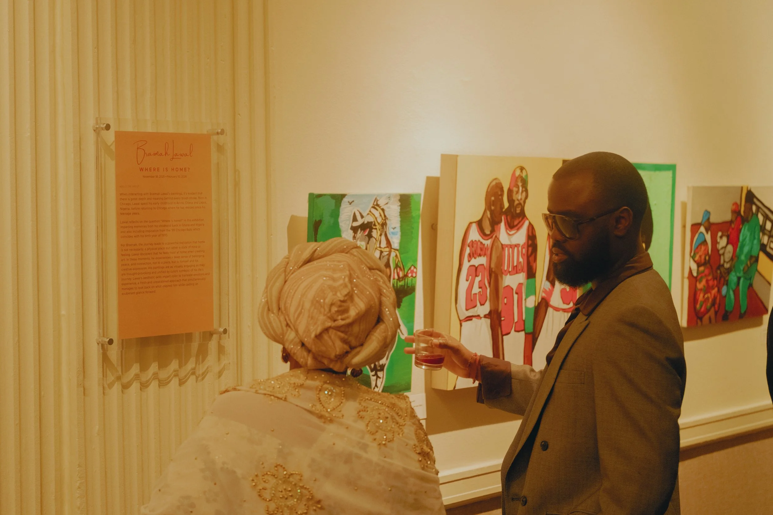 A man and woman having a conversation at an art gallery. The woman is dressed in traditional clothing with a headwrap, and the man is wearing glasses and a suit. They are looking at colorful portraits and a sign on the wall.