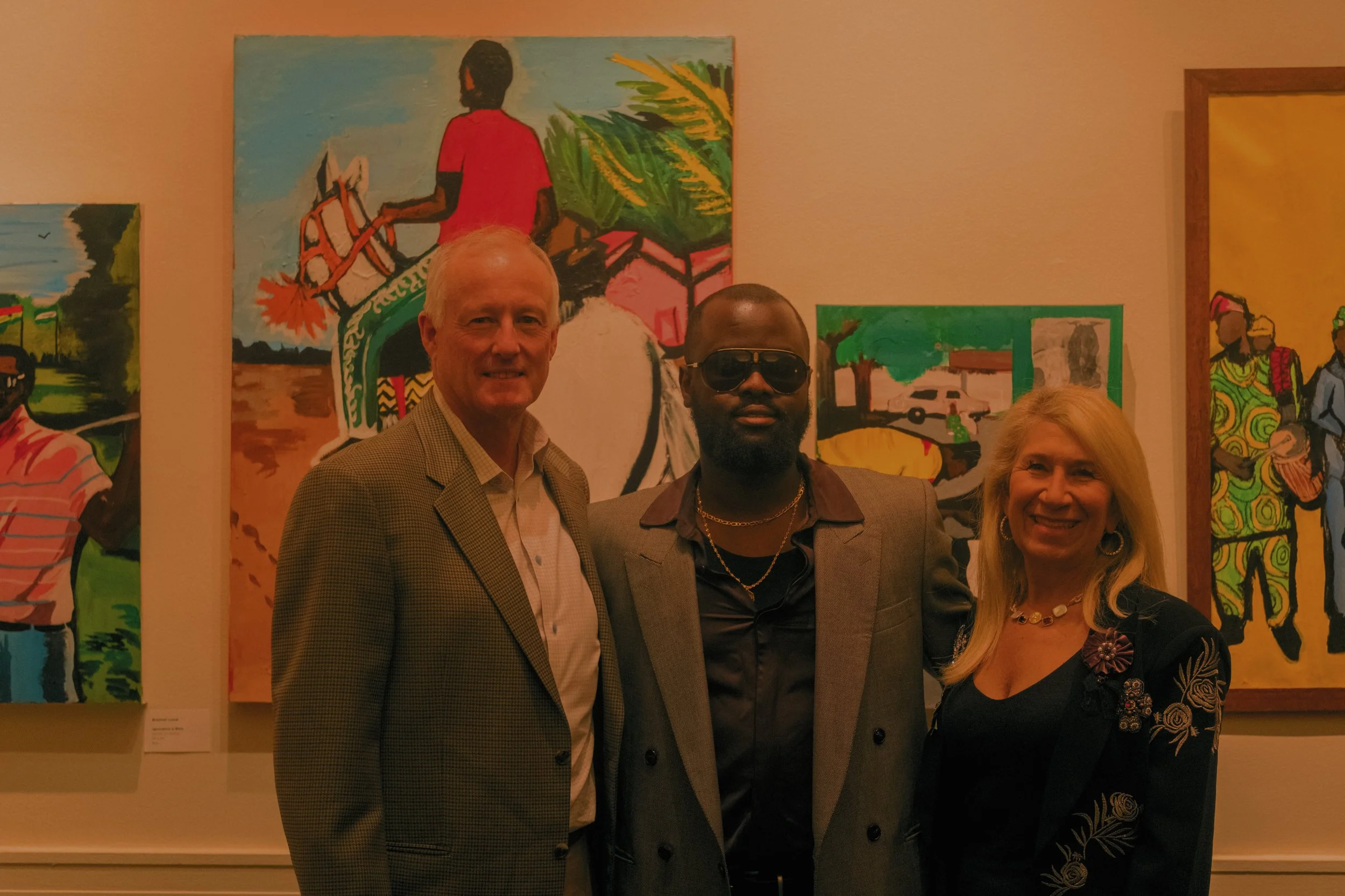Three people standing in front of colorful paintings, smiling at an art gallery or exhibition.