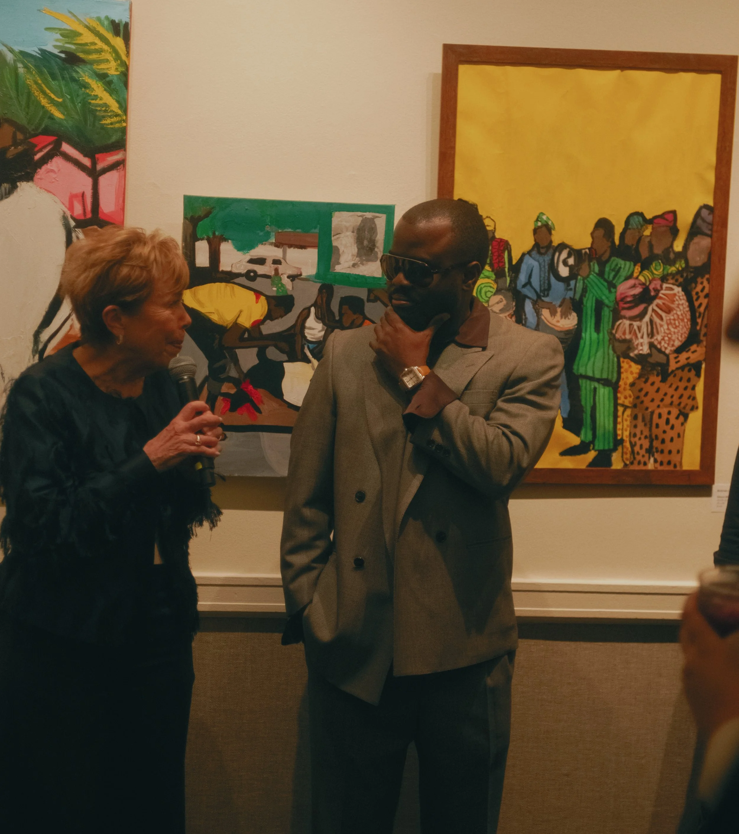 An older woman with short, light-colored hair holding a microphone and speaking to a man in a gray suit with dark sunglasses at an art gallery exhibit. There are colorful paintings of African scenes on the wall behind them.