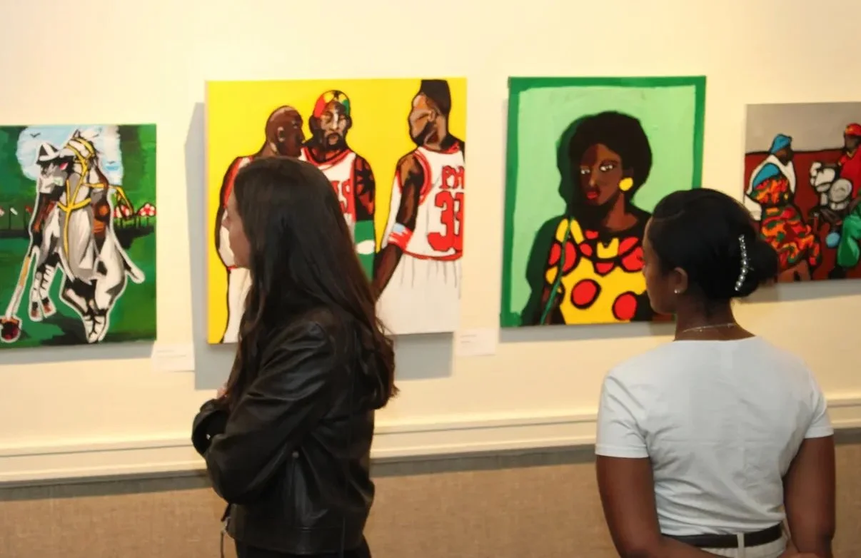 Two women viewing colorful paintings of African American figures and characters in an art gallery.