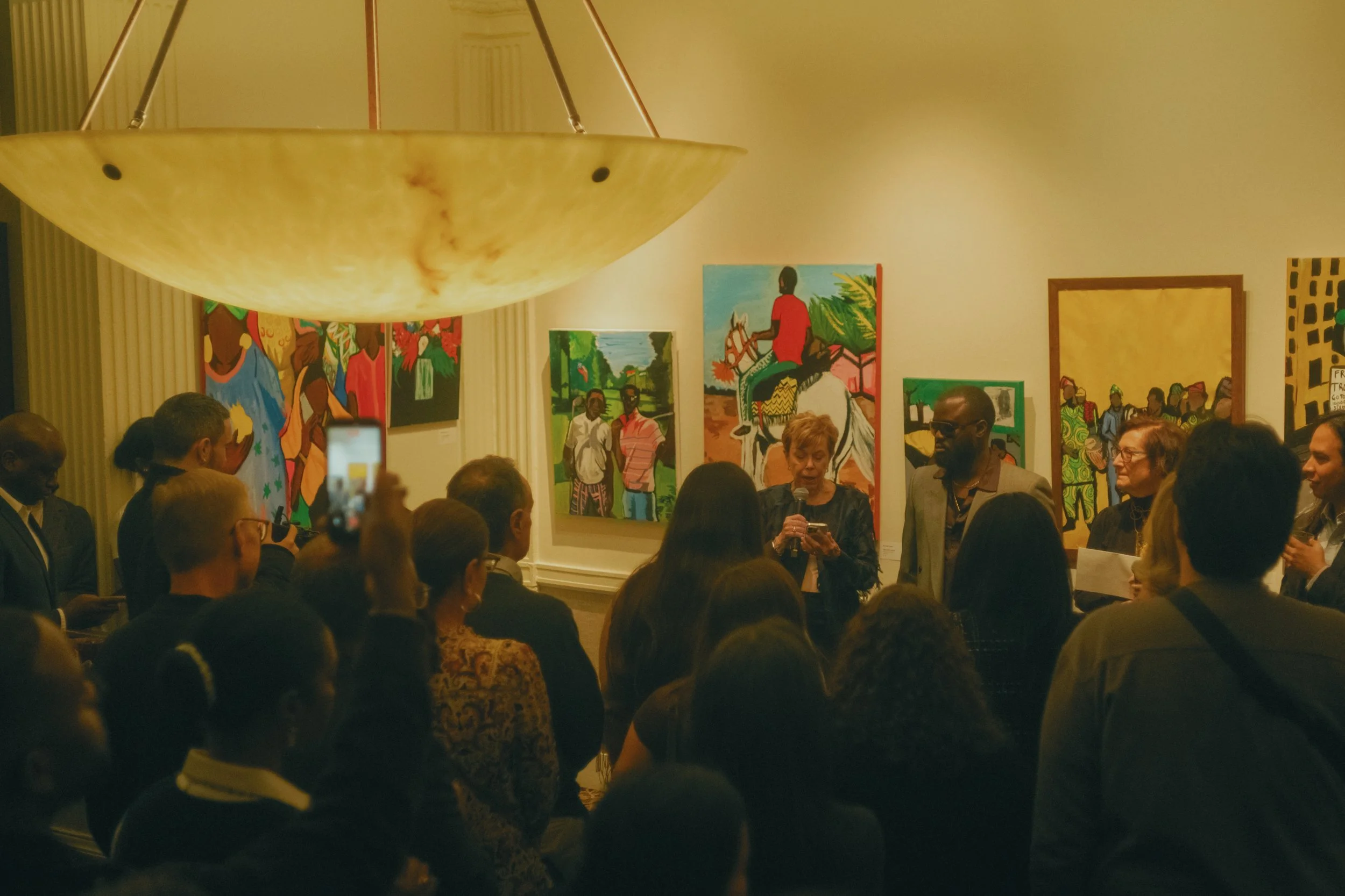 People attending an art gallery opening, listening to a woman speak with a microphone, surrounded by colorful paintings on the walls.
