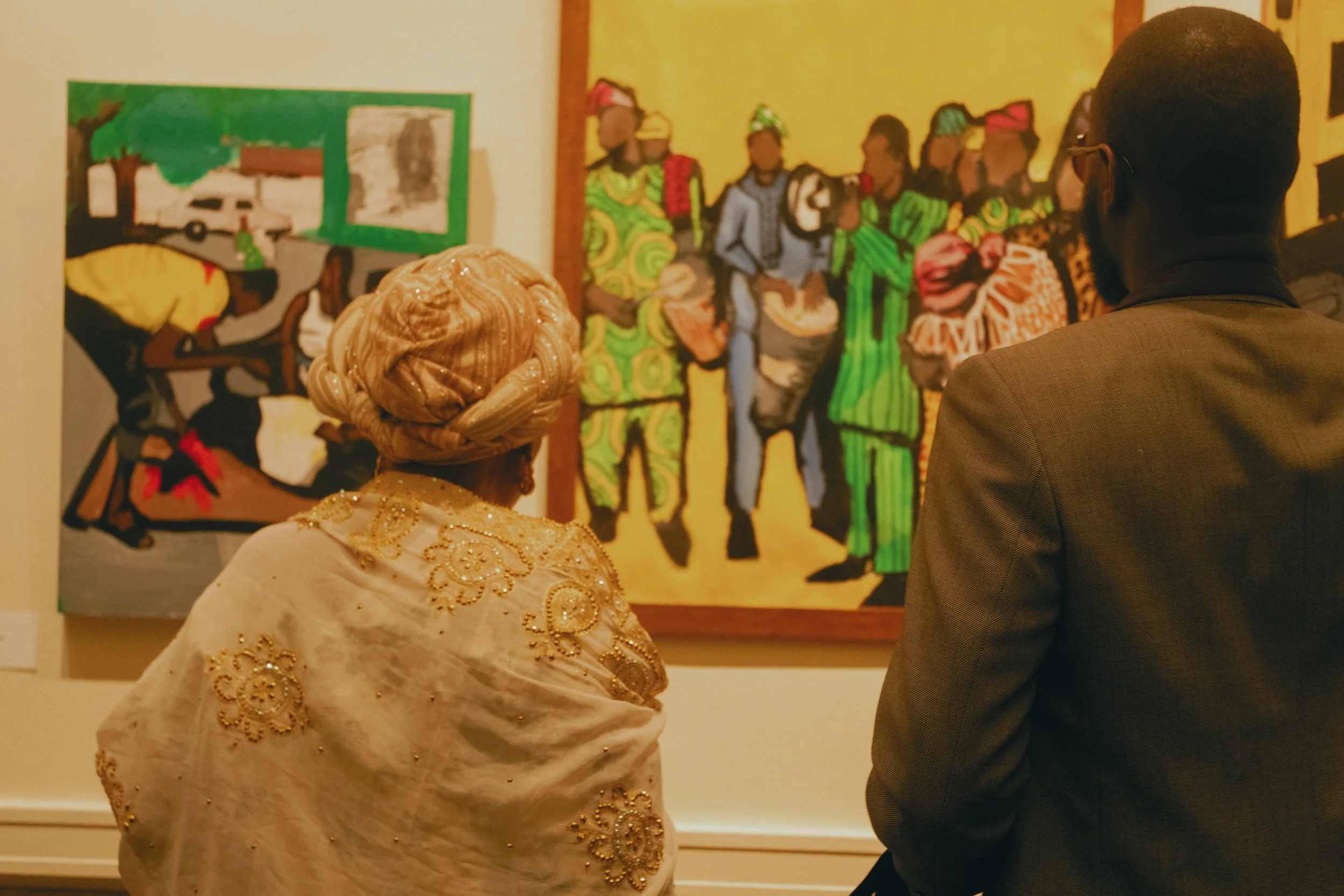 Two people observe paintings in an art gallery. One is a woman wearing a gold patterned headwrap and traditional attire, and the other is a man in a brown suit. The paintings depict African culture and scenery.