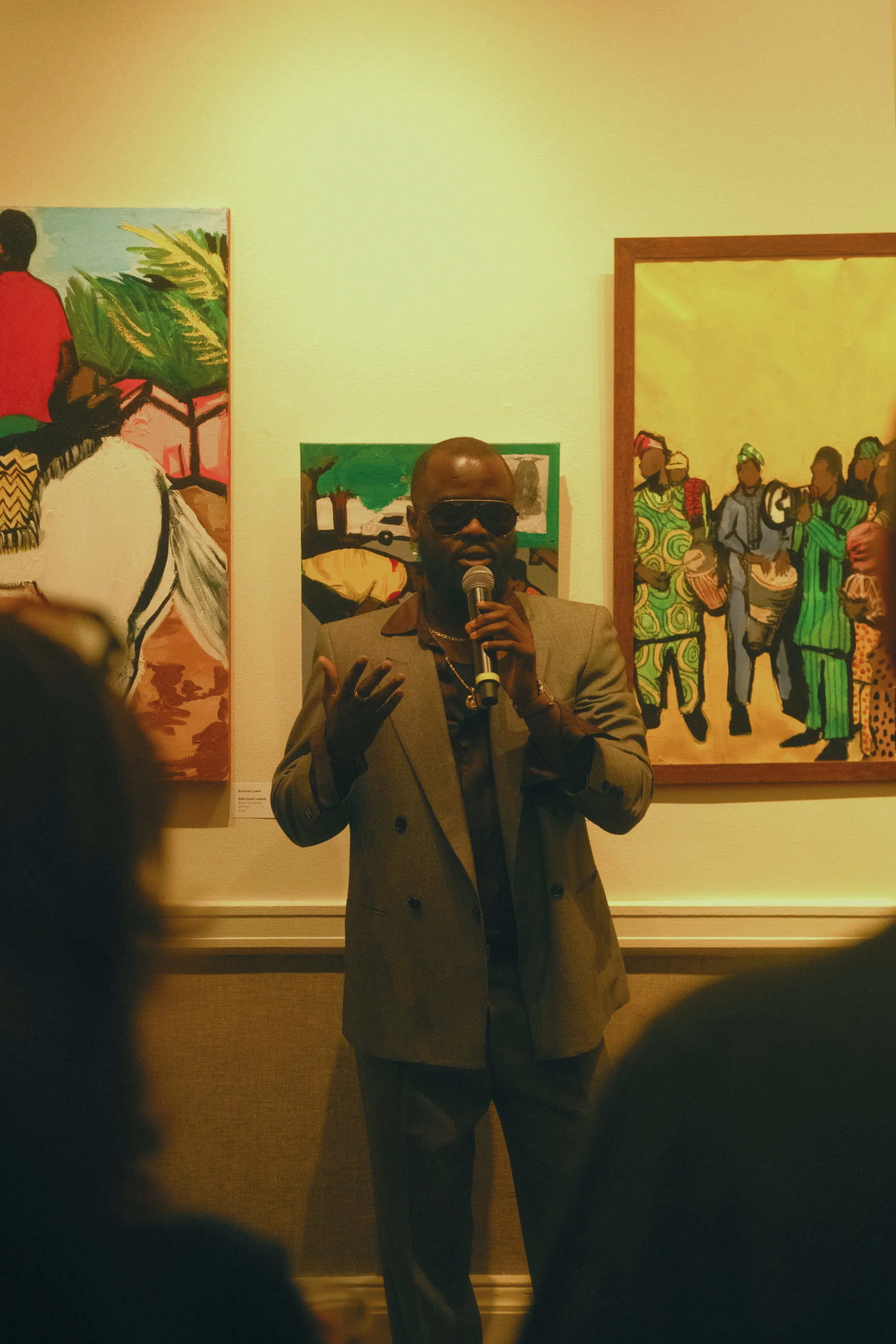 Man in a gray suit and sunglasses speaking into a microphone at an art gallery, with colorful paintings in the background.