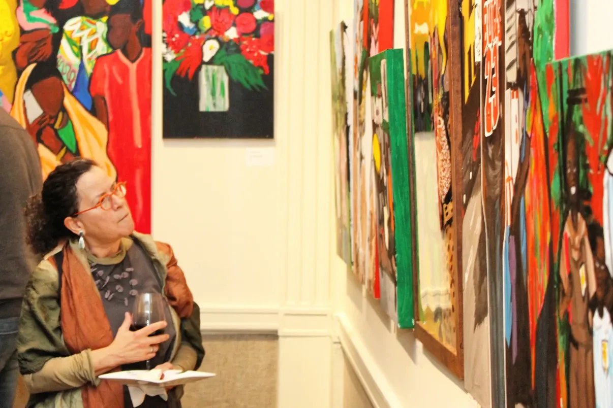 Woman with curly dark hair, red glasses, and wearing a green jacket and orange scarf, viewing colorful modern paintings at an art gallery. She holds a glass of red wine and a notebook.
