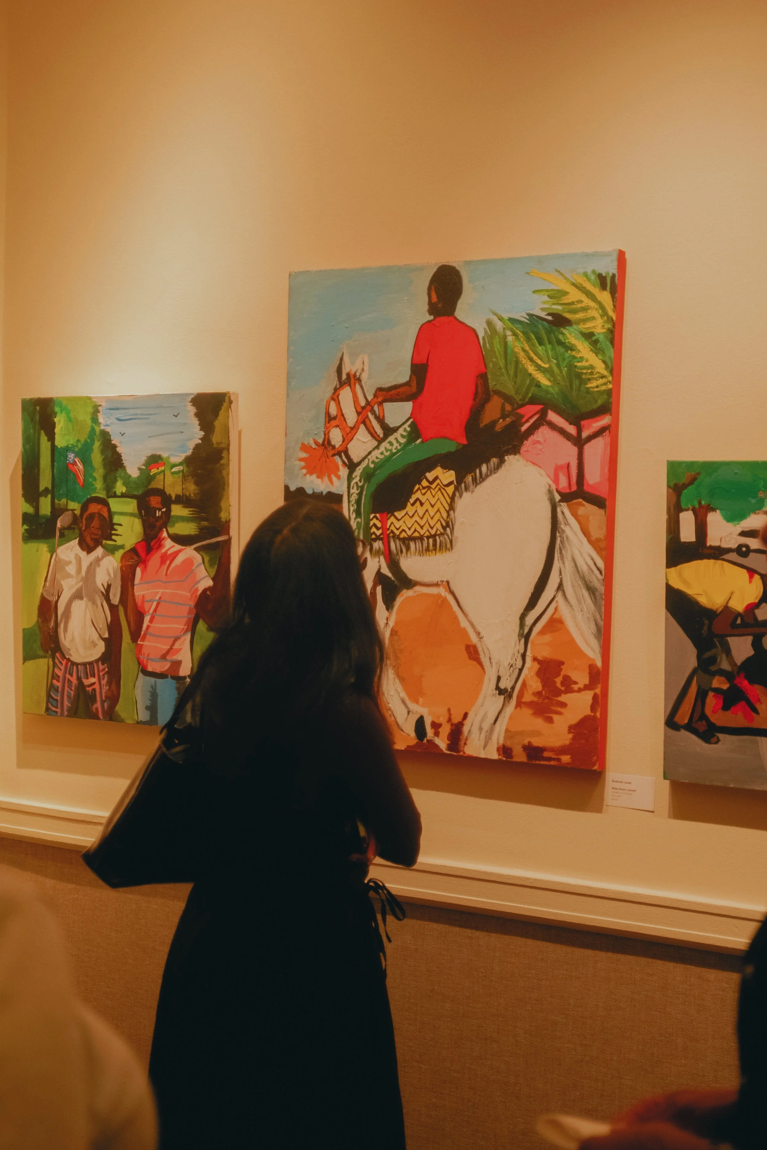 Person viewing colorful paintings in an art gallery, with one painting depicting a person on horseback and two men standing outdoors.