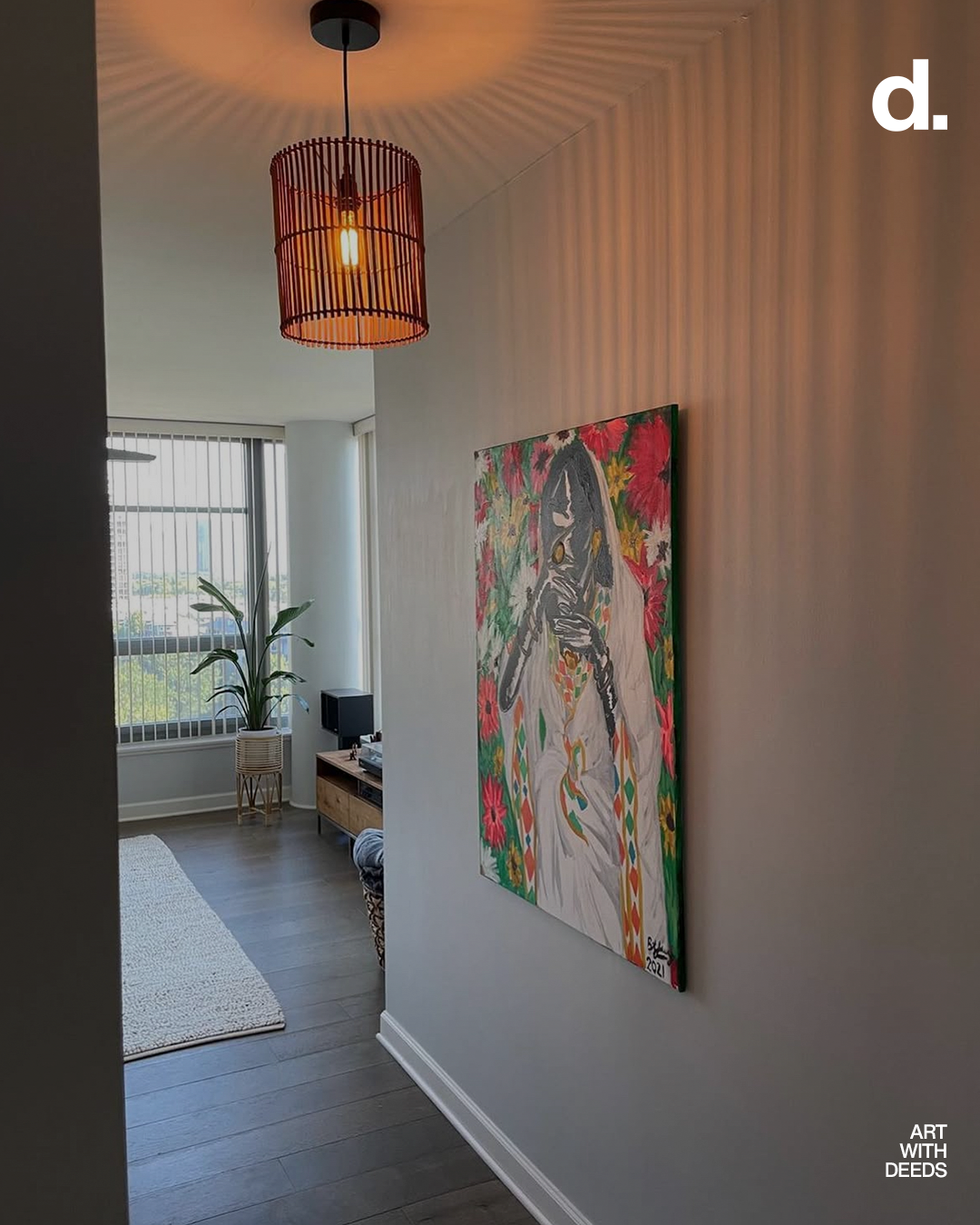 Interior of a modern apartment with a hanging orange lamp, a colorful painting on the wall, a large window with blinds, a potted plant, a small black speaker, and a TV stand.
