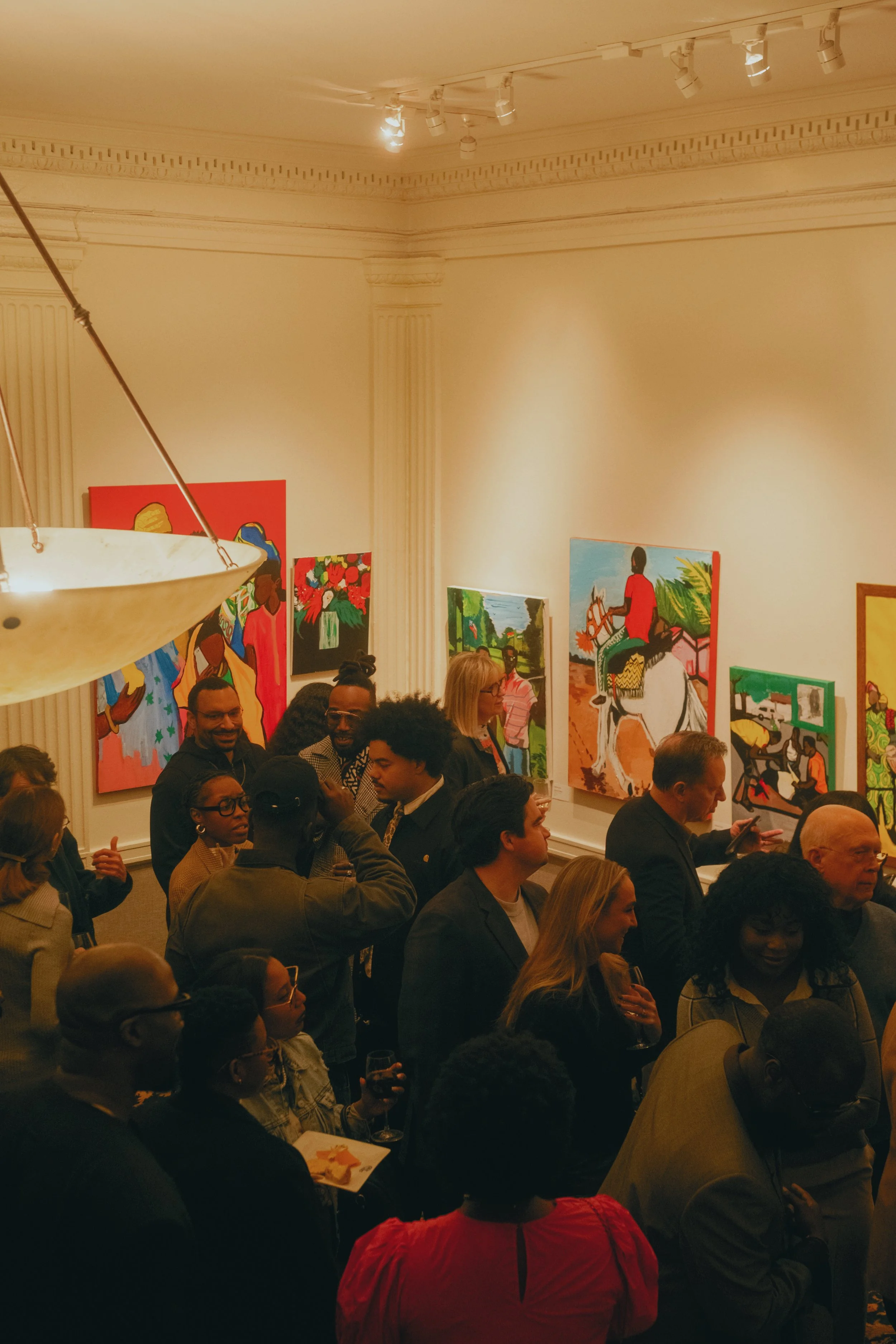 People attending an art exhibition in a gallery, viewing colorful paintings on the walls, some holding wine glasses and engaging in conversation.