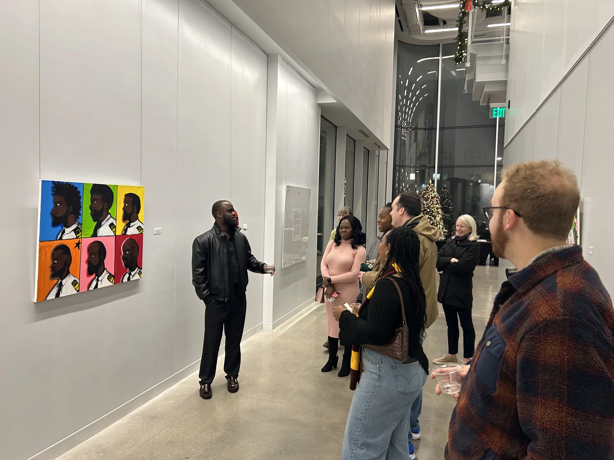 A group of people in an art gallery listening to a speaker. The artwork on the wall features colorful portraits of a man with different hairstyles and backgrounds.