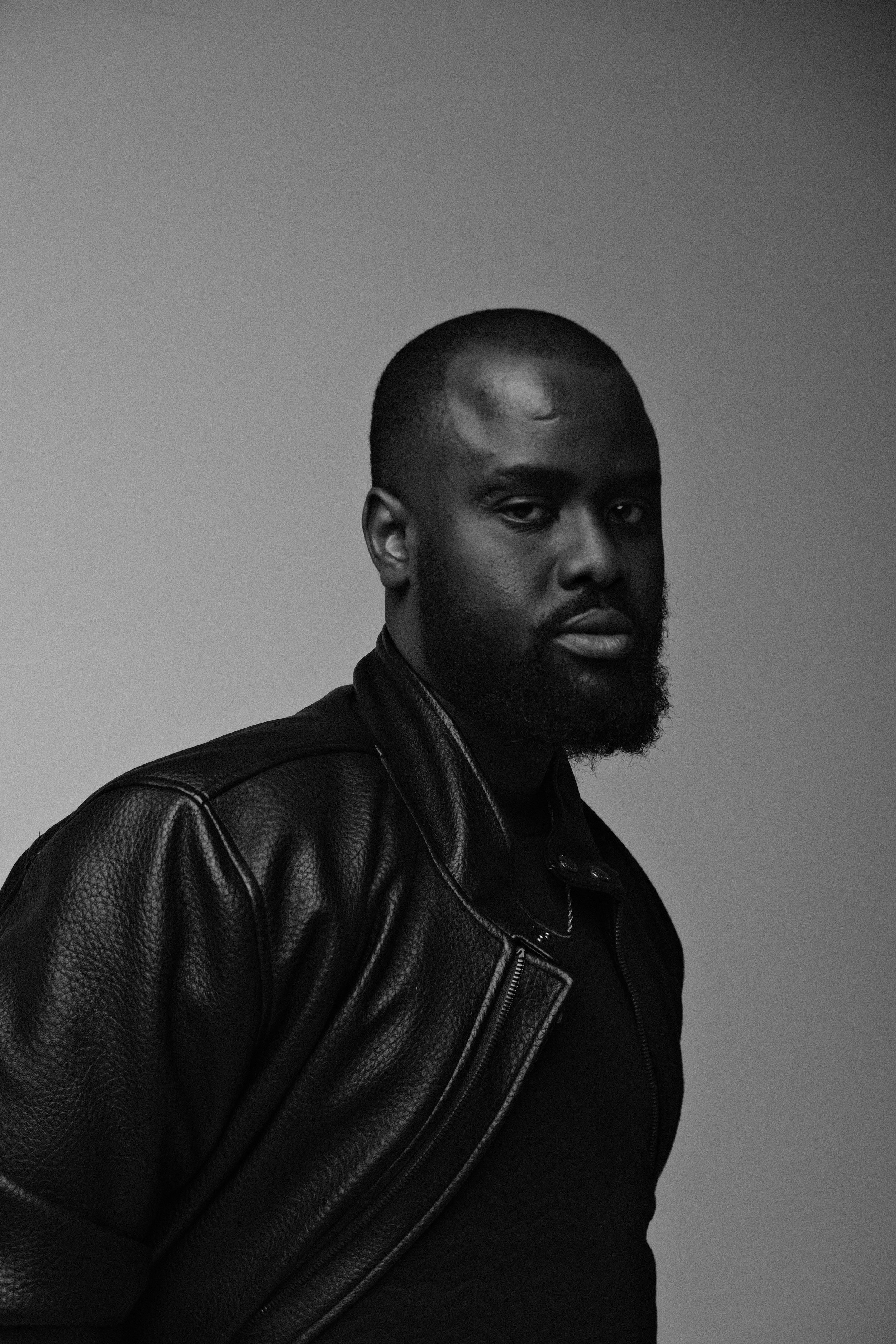 Black man with a beard wearing a black leather jacket and a black shirt, looking at the camera against a plain background.