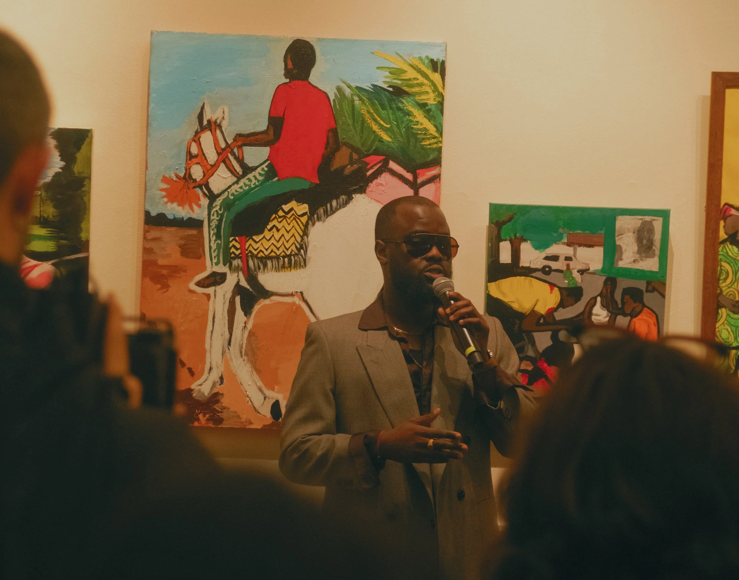 A man in a gray suit and sunglasses is speaking into a microphone at an art gallery, with colorful paintings of African scenes in the background.