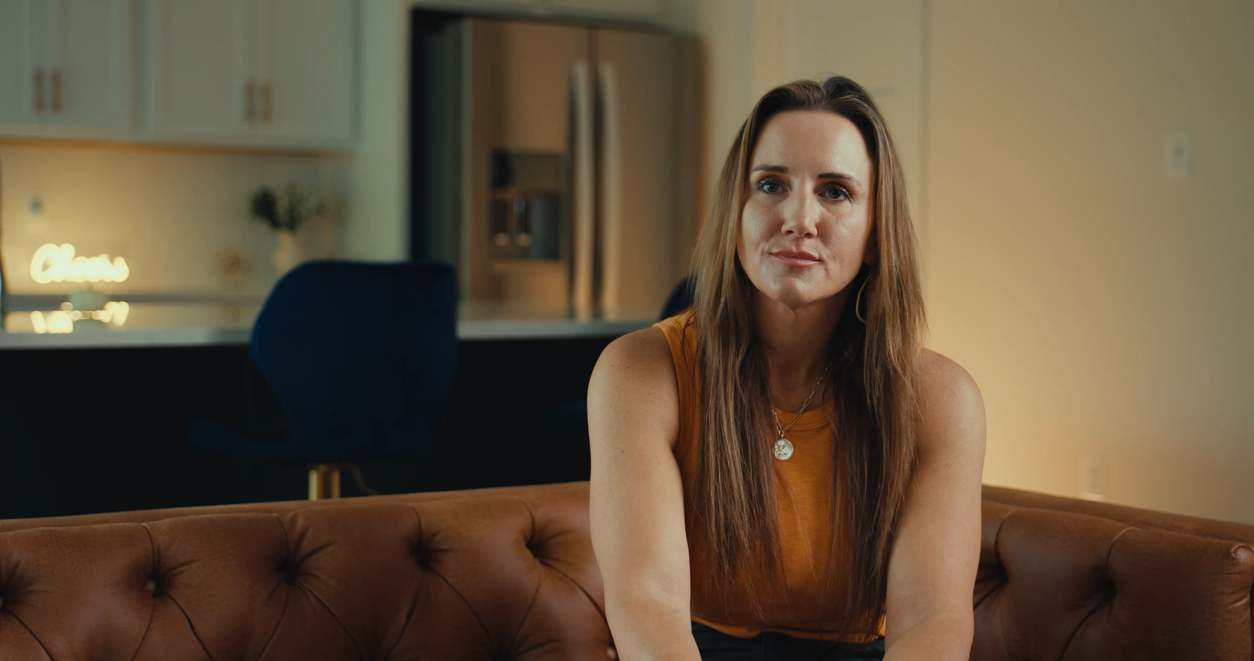 A woman with long brown hair wearing an orange top and jewelry sitting on a brown leather couch in a modern living room.