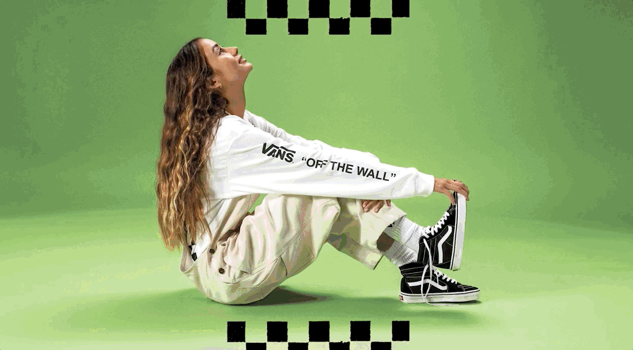 A woman with long, wavy hair sitting on the floor in a studio with a green background, wearing a white Vans sweatshirt, light-colored pants, and black Vans sneakers, looking upward.