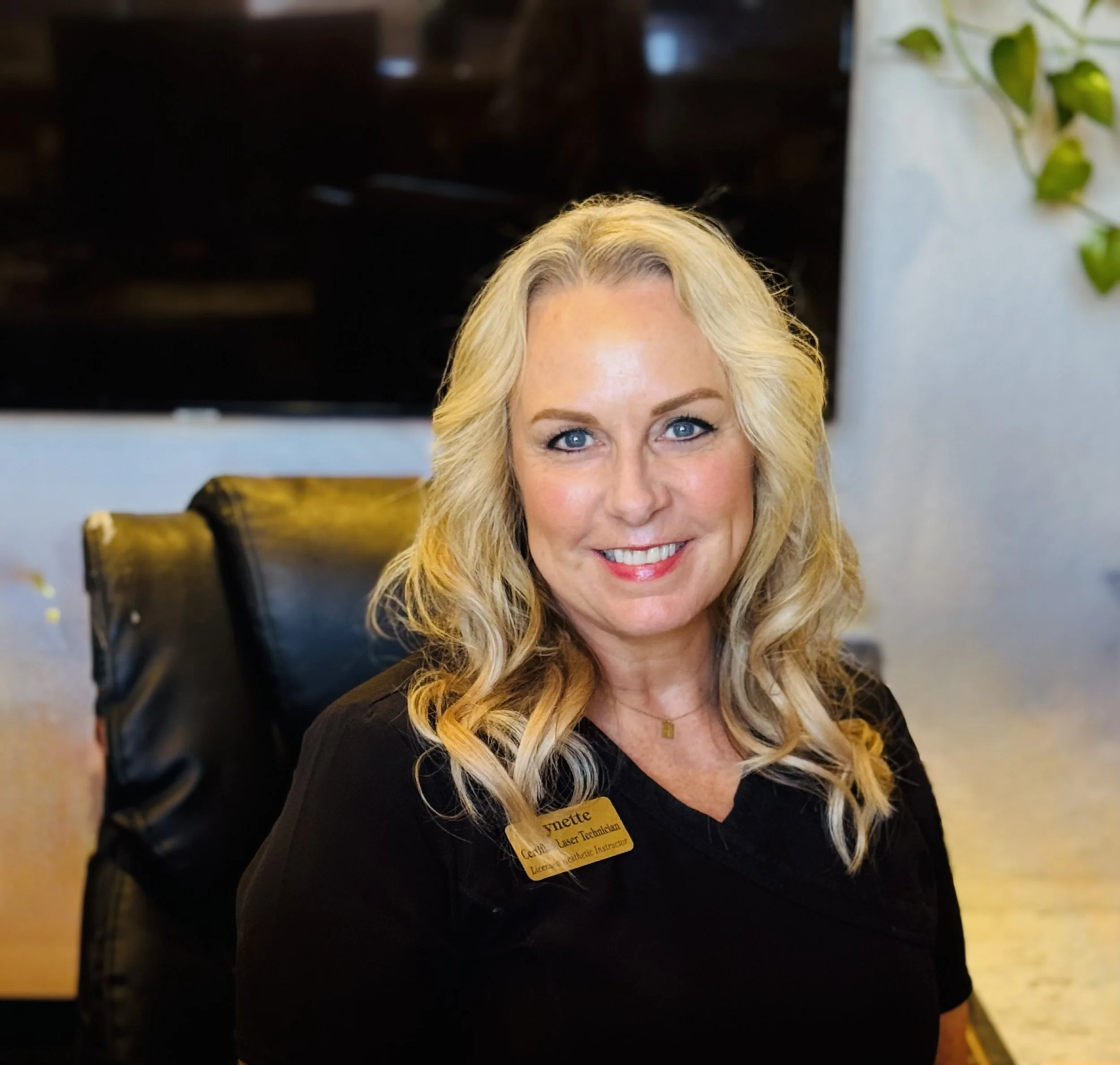 A smiling blonde woman with blue eyes sitting in a black leather chair, wearing a black top and a gold name tag, in a well-lit indoor setting with indoor plants in the background.
