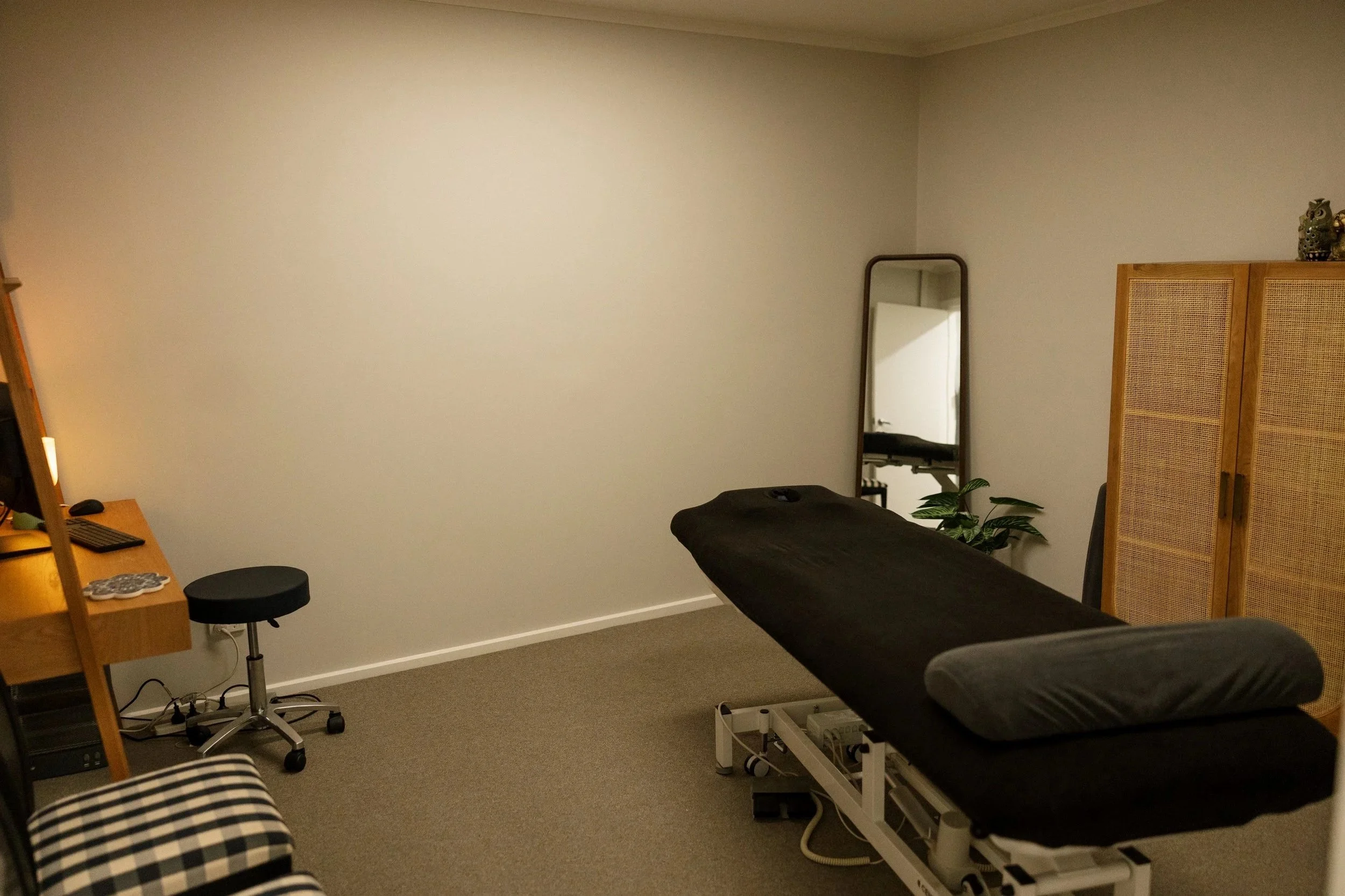 A massage room with a massage table covered in black sheets, an adjustable pillow, a mirror, a small potted plant, a wooden cabinet, and a computer setup with a stool, in a room with beige carpet and plain white walls.