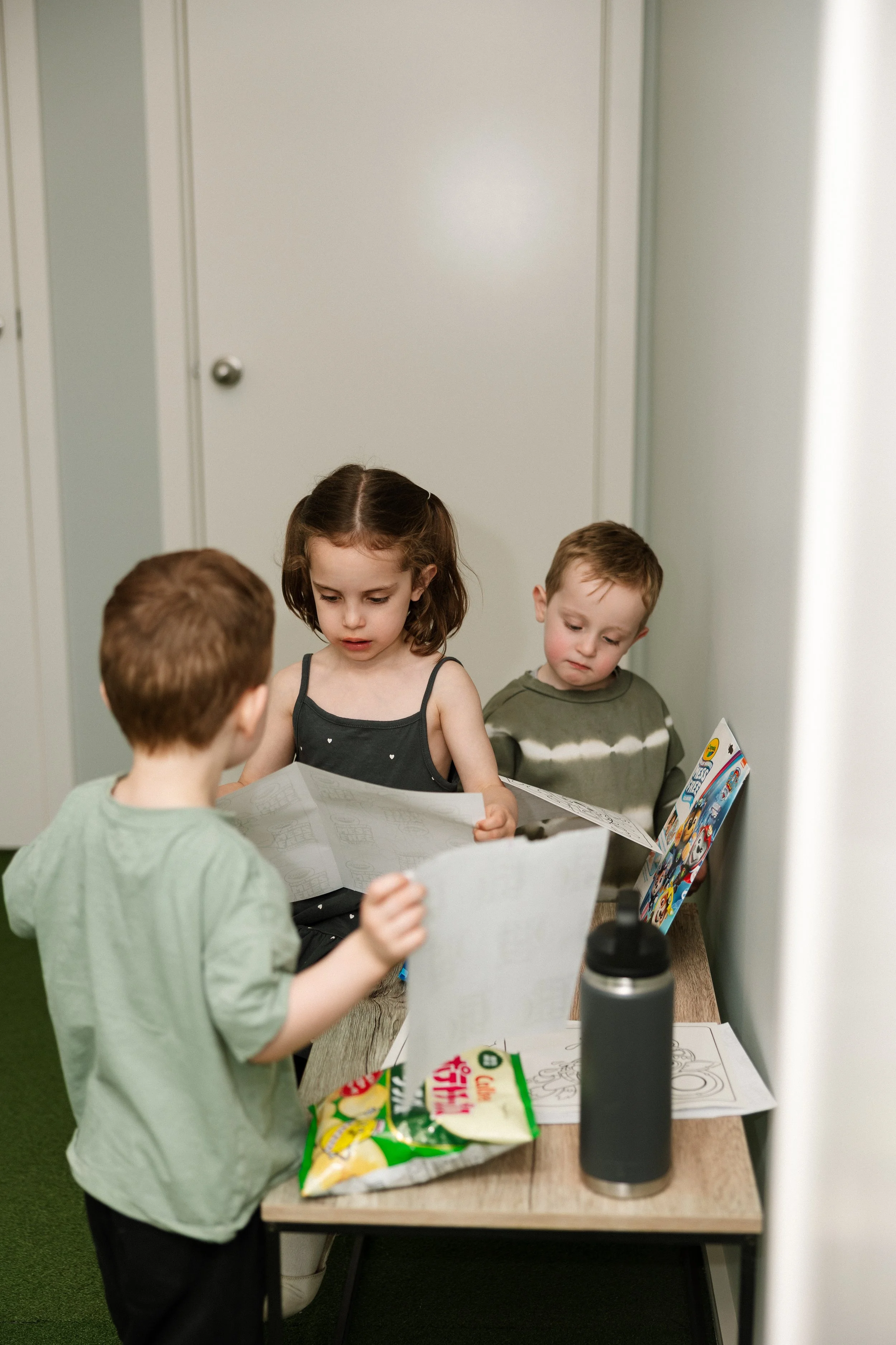 Three young children looking at coloring books and papers on a small wooden table with a snack bag and a black water bottle, in a room with plain light-colored walls and a closed door.