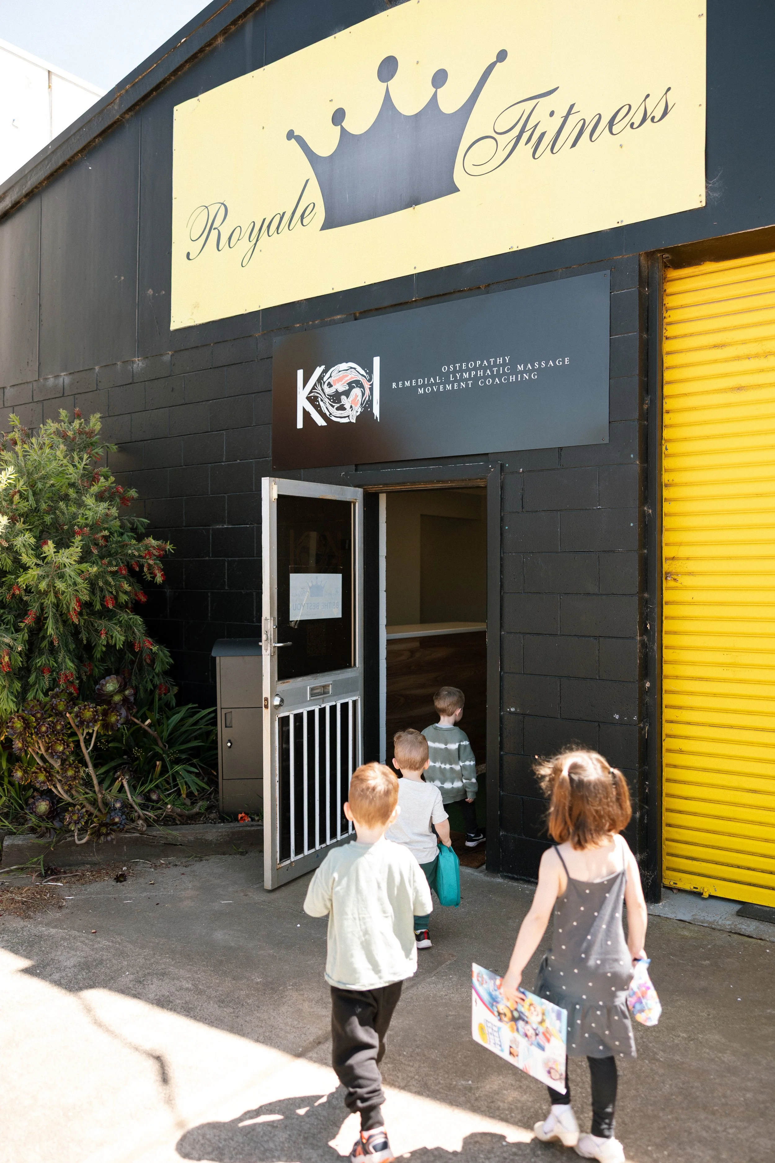 Four children walking into a black building with signs that read 'Royal Fitness' and 'K.O.I Osteopathy, Remedial, Lymphatic Massage, Movement Coaching.'