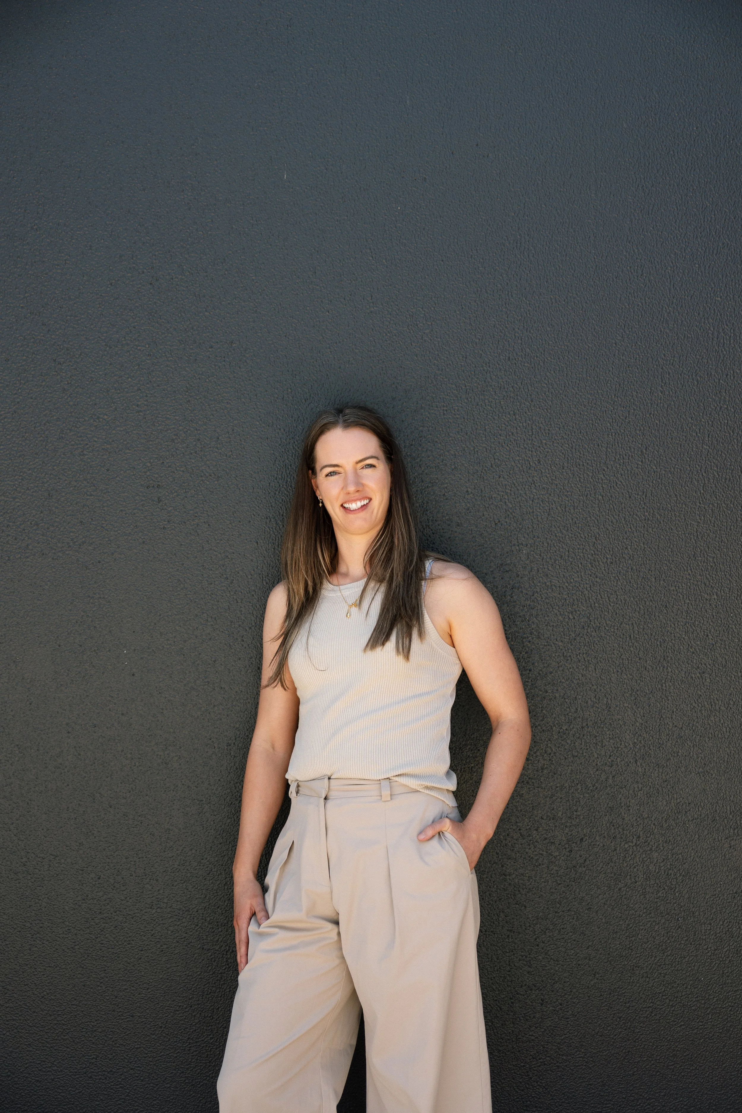 A smiling woman with long brown hair standing in front of a dark gray textured wall, wearing a sleeveless beige top and loose beige pants with one hand in her pocket.