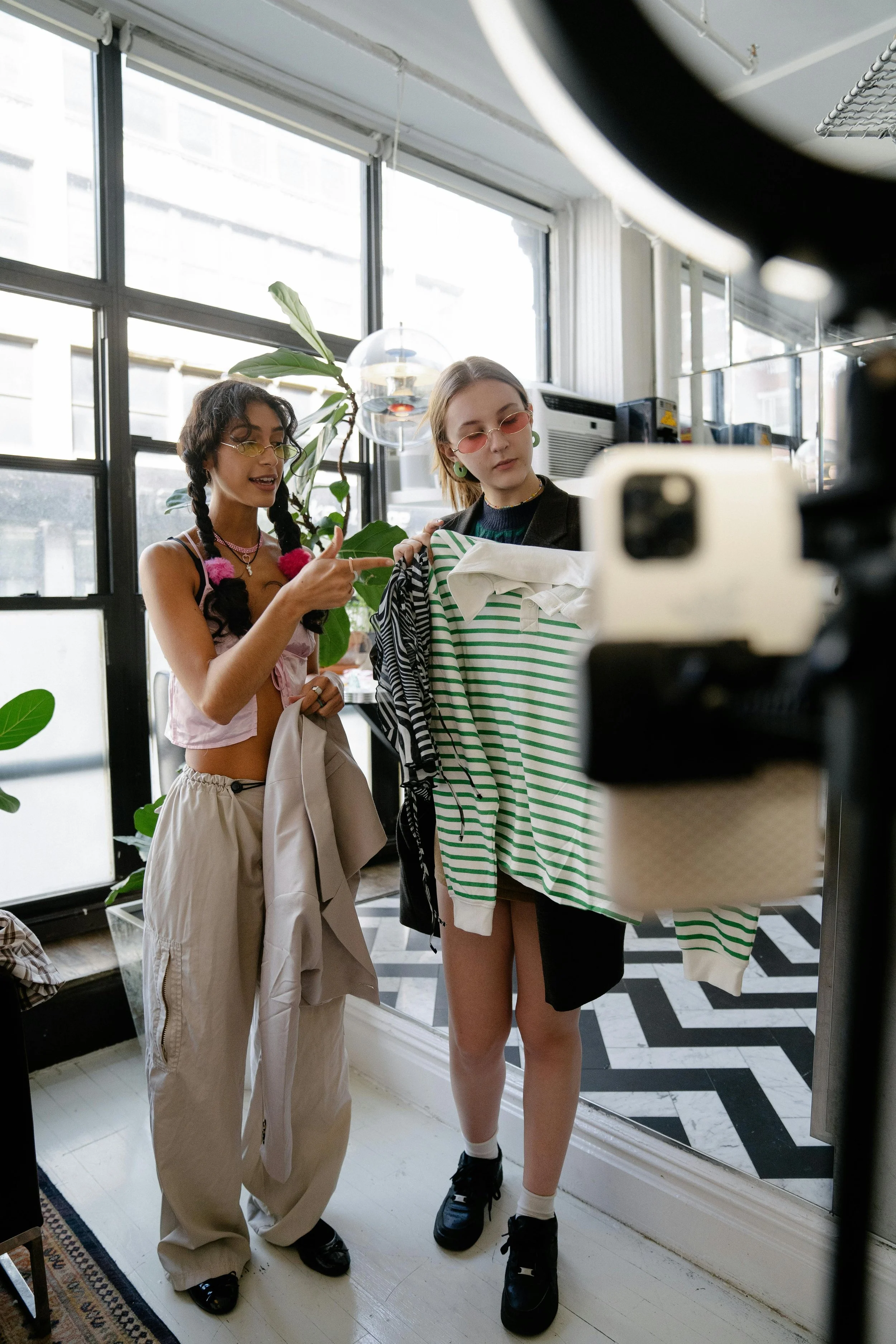 Two young women shopping for clothes, one is pointing at the clothing while the other looks at a hanger with a green and white striped sweater. They are inside a well-lit store with large windows and modern decor.