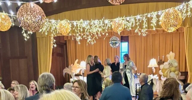 People socializing at a party with gold and white decorations, hanging lights, and balloons.