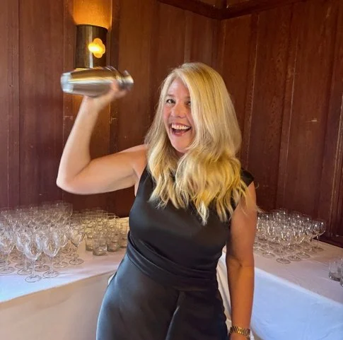 A woman with blonde hair wearing a black dress, smiling and holding a cocktail shaker above her head in a room with wooden walls and a table with many empty glasses.