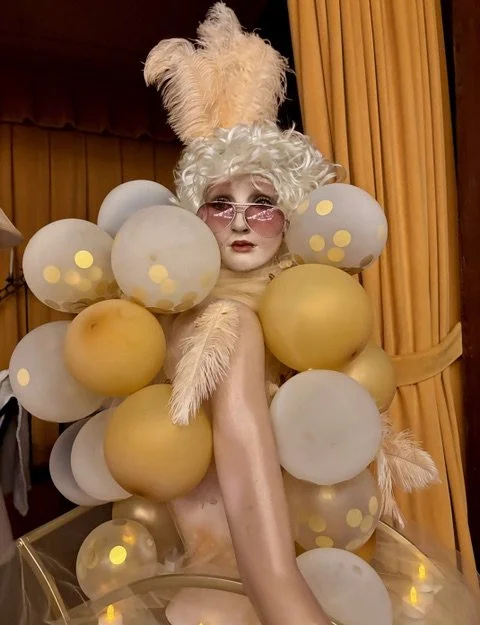 Person dressed in an elaborate costume with large white and yellow balls, fluffy feathers, and pink sunglasses, posing in front of orange curtains.