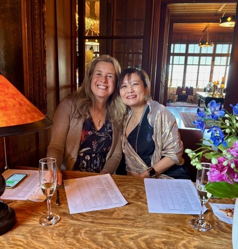 Two women smiling at a table with papers and drinks, in a warmly lit room with large windows and wooden decor.
