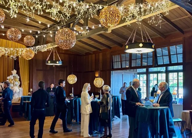 People socializing at a decorated indoor event space with string lights, hanging lanterns, and large windows.