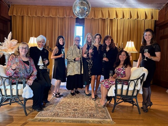 Group of nine women in a warmly decorated room with curtains, lamps, and a small window, holding glasses of champagne or wine, dressed in formal and semi-formal attire, some seated and others standing.