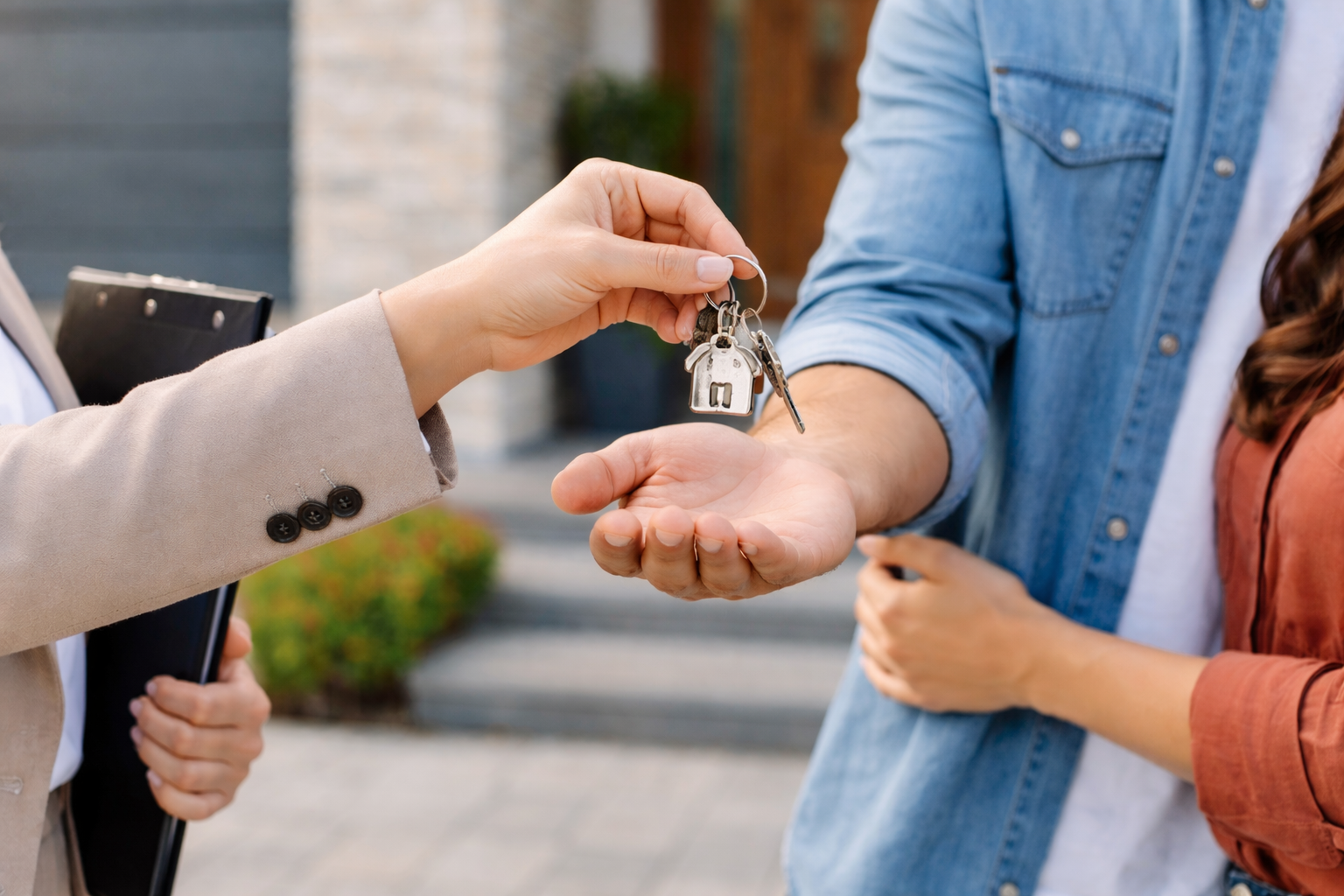 A person in a beige blazer handing over house keys to a couple, outside in front of a building.