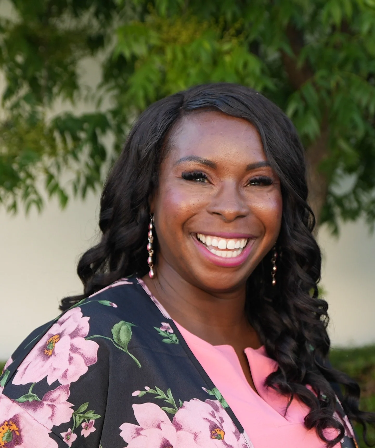 A woman with long, curly black hair, smiling outdoors in front of green trees. She is wearing a pink top with a black floral blazer and pink earrings.