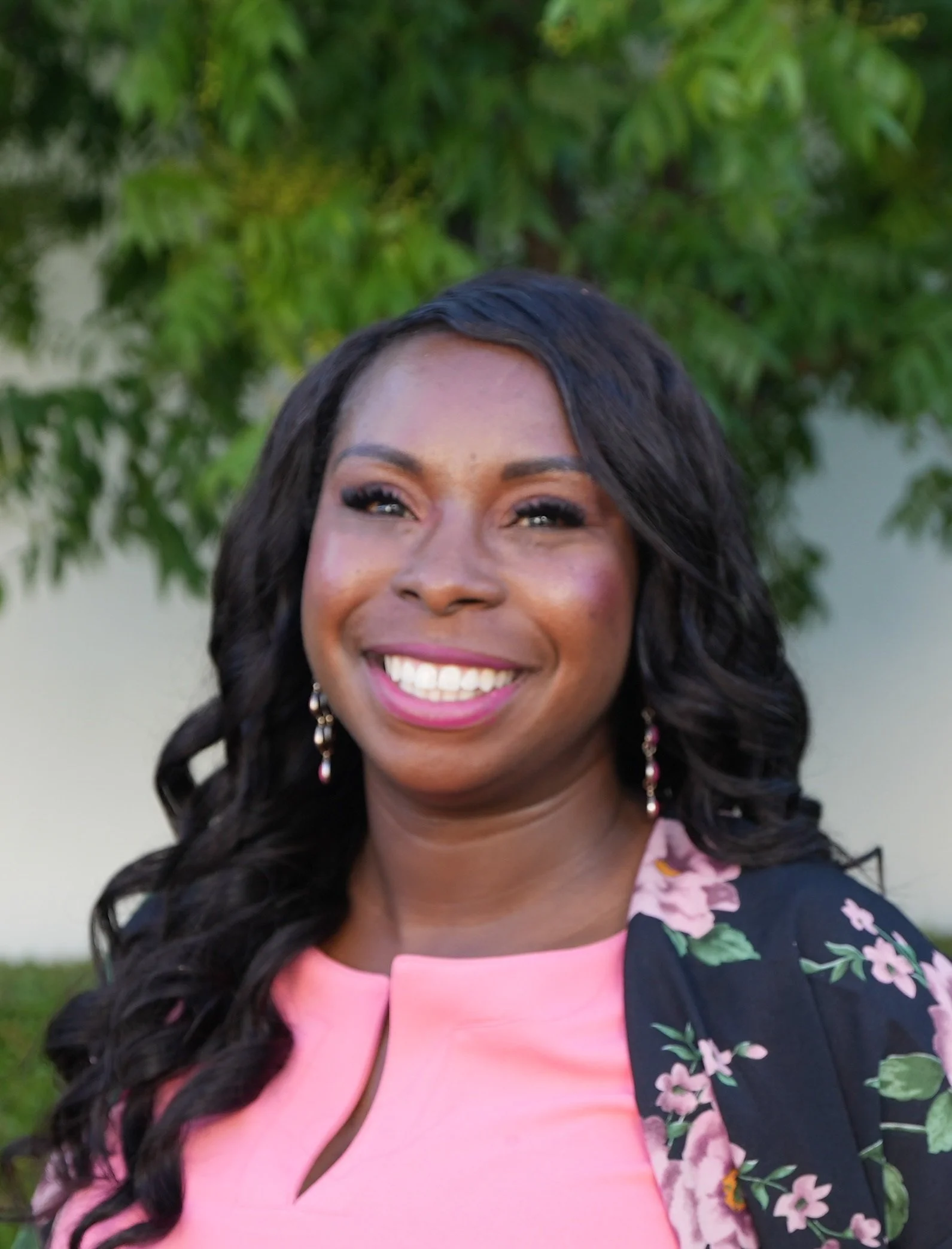 A smiling woman with dark curly hair, wearing a pink top and floral blazer with earrings, standing outdoors with green trees in the background.