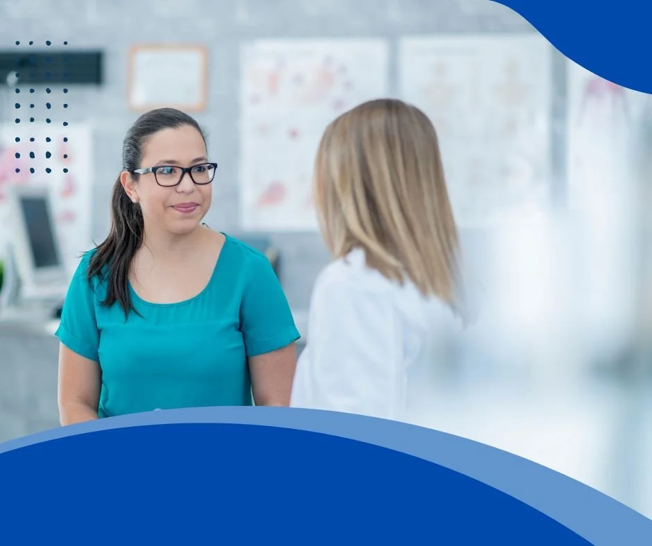 A woman in glasses and a teal shirt talking to another woman in a white coat in a clinical setting, with medical charts and computers in the background.