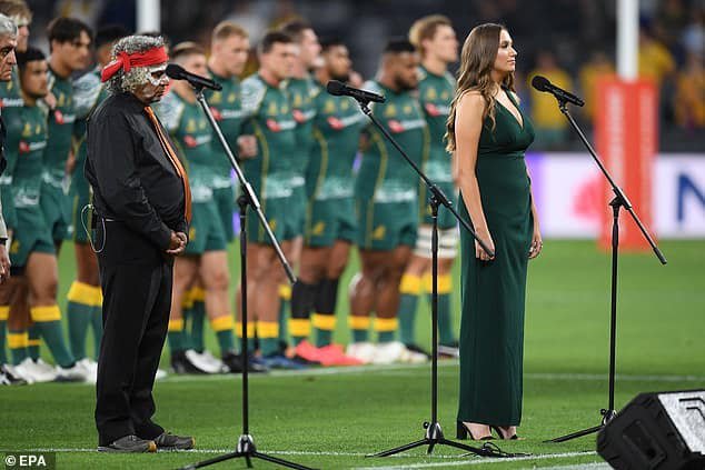 A woman in a green dress and an older man with a gray beard, glasses, and a red headband stand at microphones during a ceremony on a sports field, with a line of athletes in green uniforms standing behind them.