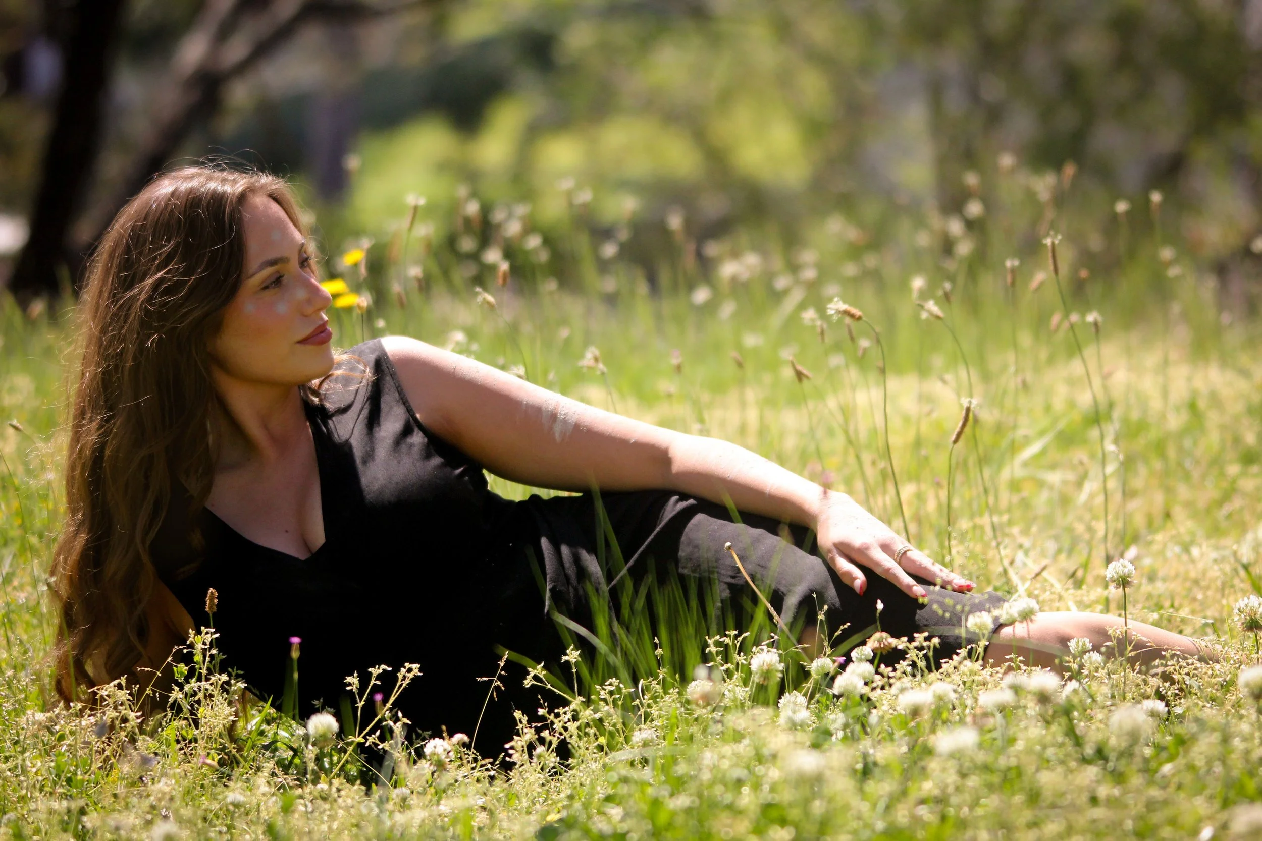 A woman in a black dress lying in a field of grass and white wildflowers on a sunny day.