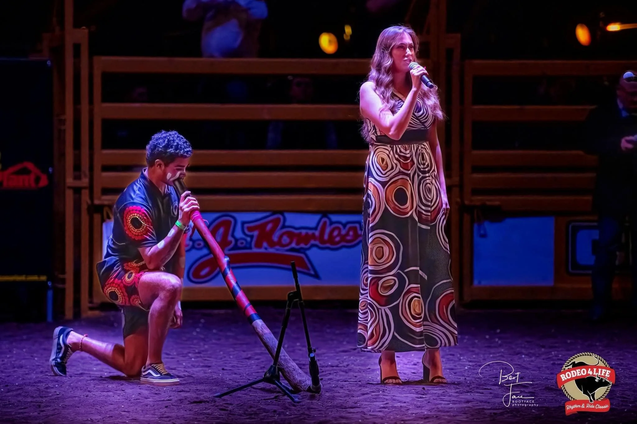A woman in a long patterned dress singing into a microphone on stage, while a man in a Native American-inspired shirt and shorts kneels holding a decorated wooden flute. Stage lighting is visible in the background.