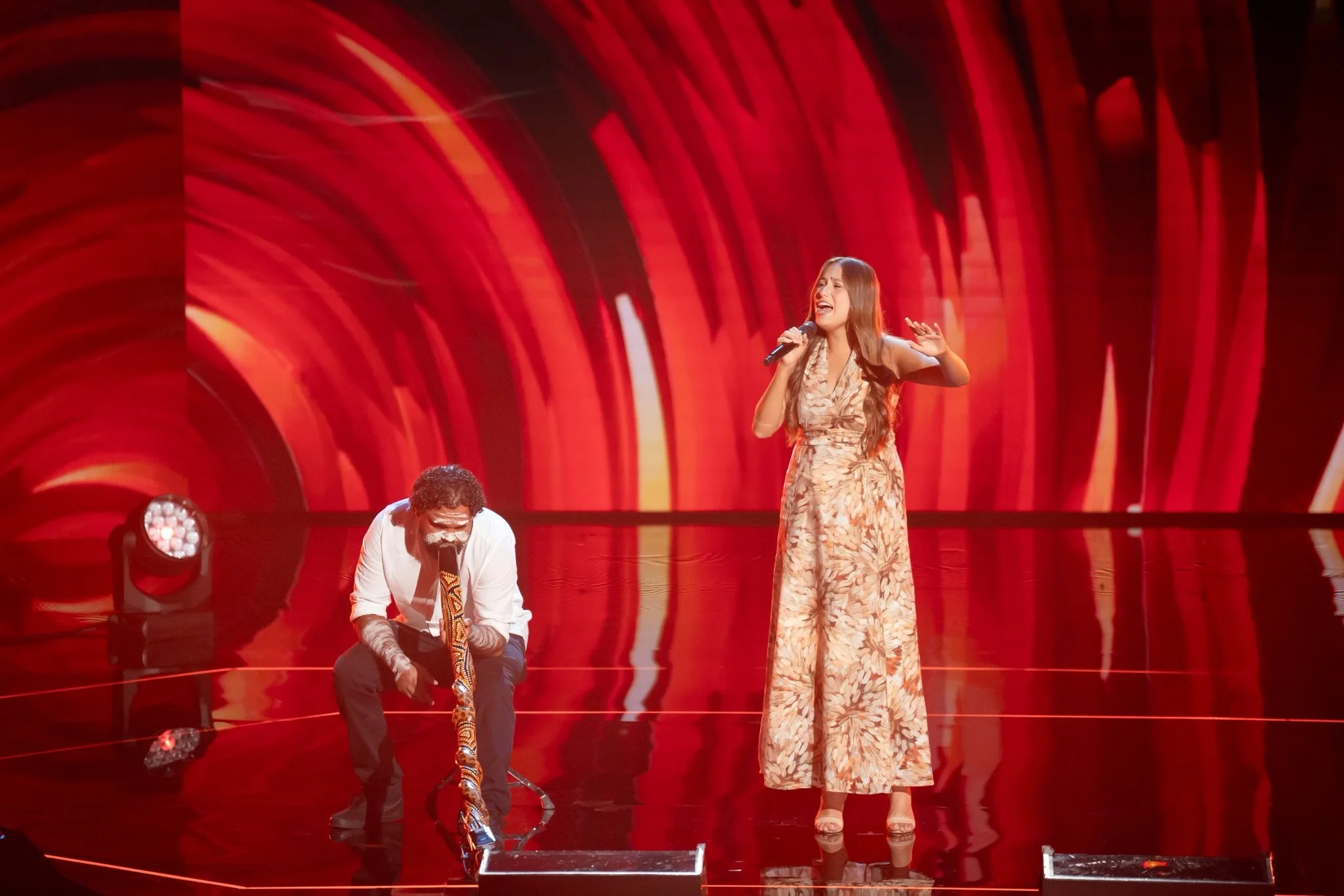 A woman singing on stage with a man playing a snake in front of her, set against a red and orange swirling background, with stage lights shining.