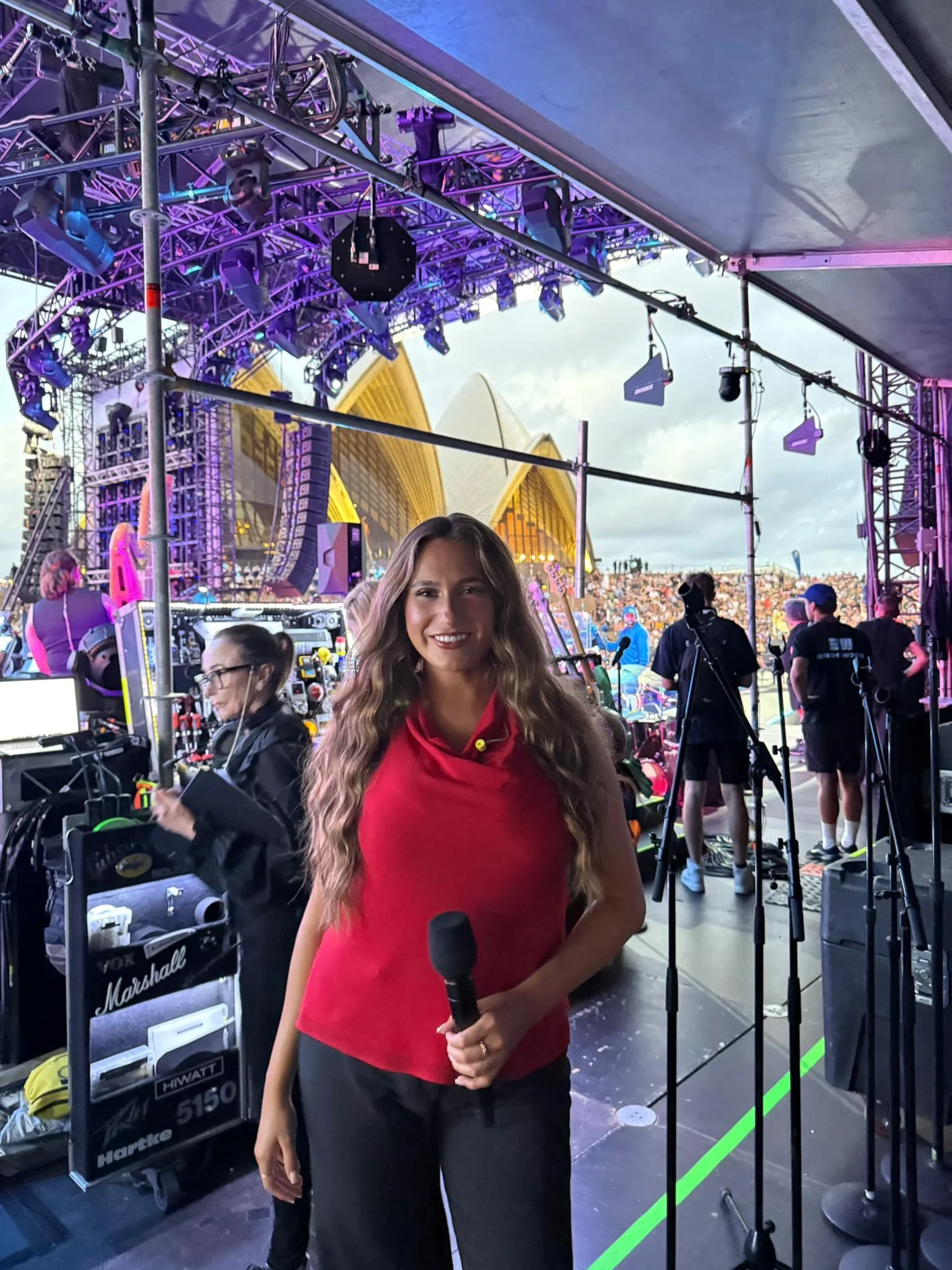 A woman holding a microphone on stage at an outdoor concert, with stage equipment, crew members, and the Sydney Opera House in the background.