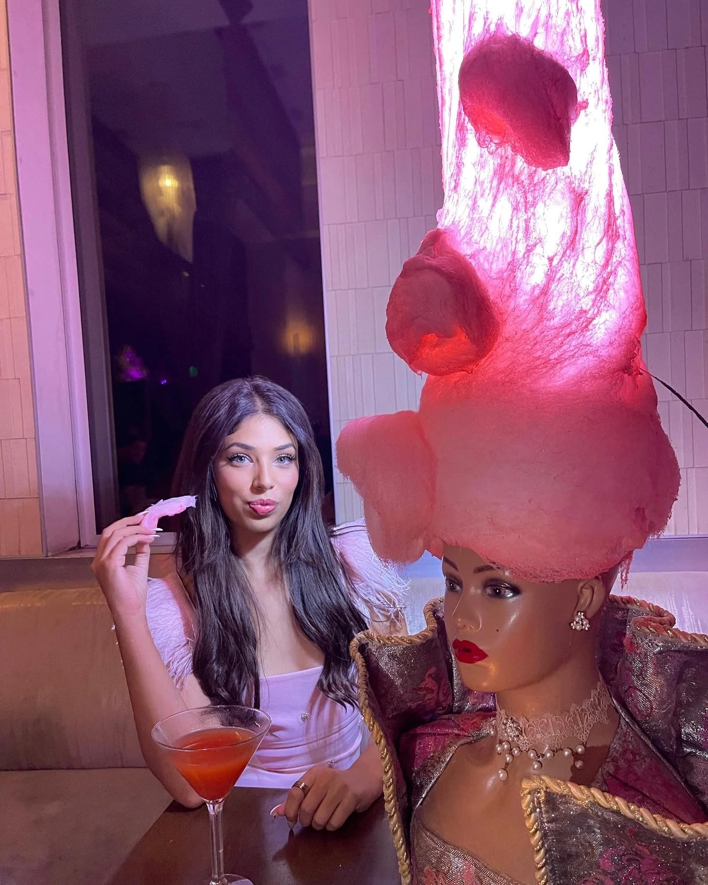 A woman with long dark hair sitting at a table in a pink-lit setting, holding a pink object, with a mannequin or costume figure wearing a large pink headdress, jewelry, and makeup in front of her.